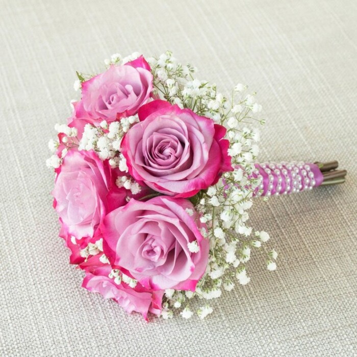 Five prominent roses form the heart of this elegant, round bridal bouquet, designed in Highgate to evoke the seamless blend of tradition and modern artistry beloved in the area. Each rose showcases a stunning gradient, with its outer petals suffused in vivid fuchsia pink that dissolves gently into a lavender centre-turning each bloom into a miniature masterpiece. Accenting the roses, fine sprigs of white baby's breath float in airy clusters, imparting volume and a soft, cloud-like quality to the bouquet. Attention to detail shines in the ribbon-wrapped handle: luxuriously smooth and deep purple, expertly spiralled with lustrous white pearls for a classic touch reminiscent of weddings in Highgate's historic venues. The bouquet's neat, hand-tied stem ends-rich in earthy green-peek beneath the ribbon, hinting at the freshness and local provenance cherished by the community. All of this is presented atop a subtly textured oatmeal fabric, echoing the calming, natural tones of Highgate Wood and surrounding green spaces. The overhead perspective captures both the voluptuous shape and delicate artistry, making the bouquet perfect for bridesmaids, flower girls, or those seeking inspiration for sophisticated ceremonies and celebrations unique to North London.