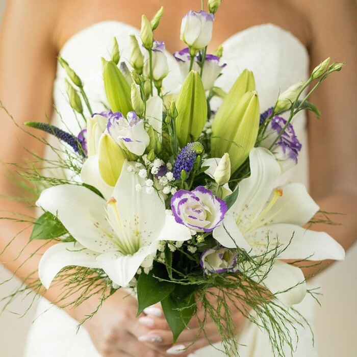 A breath of botanical artistry sweeps through this close-up of a bridal bouquet held lovingly by a bride in elegant Highgate surroundings. The ensemble features a medley of grand, creamy white Oriental lilies-some fully open, others peeking from still-tight buds-each flaunting soft spotting, luminous petals, and prominent golden stamens. Interlaced throughout are plush white lisianthus, their ruffled blooms tinged with lilac at the margins, their forms echoing the classic yet creative floral displays often seen around Highgate's Village and Chorleywood woodland walks. Tall, deep purple veronica rise from within, sending vertical accents through the white and lavender haze, while ethereal sprays of baby's breath scatter across the arrangement, giving a magical, cloudlike effect. The lush greenery, including glossy lily leaves and feathery asparagus fern, wraps around the bouquet, providing organic abundance and layered depth. Light from a window spills over the bride's manicured hands-nails painted in a soft pink-while the folds of her white gown blur into a glowing backdrop, highlighting the bouquet's artistic form. Like a living memento of a Highgate wedding morning, this bouquet marries tradition, contemporary flair, and the subtle fragrance of celebration, ideal for those seeking local floral inspiration.