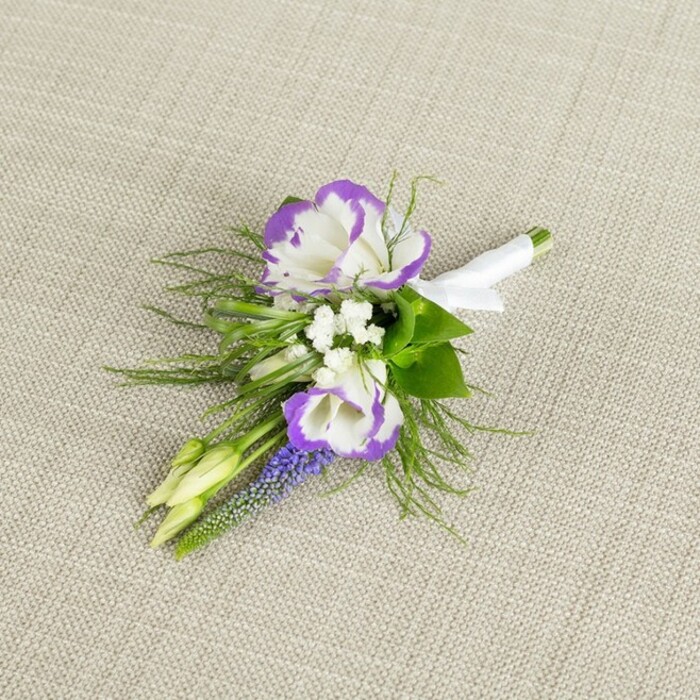 A graceful, hand-tied boutonniere is shown resting on a softly textured light beige fabric, the weave suggesting natural linen and lending a warm, understated backdrop that lets every flower detail be clearly read. Dominating the little cluster are two open lisianthus blooms, their petals a clean, luminous white with finely ruffled edges kissed by a deep, saturated purple, giving each flower a painterly, two-tone effect. Tucked around them are tiny white filler flowers, akin to baby's breath, that float between the larger blossoms like small stars, cushioning the composition with airy softness. Running vertically through the arrangement is a slender stem of muscari-style flowers, each minuscule bell a rich purple-blue, adding depth of colour and a slightly wild, woodland note. Fine, feathery greenery, reminiscent of asparagus fern, mingles with firmer green leaves at the base, creating a natural, garden-picked feel, as though these stems had just been gathered from a Highgate back garden or the edges of Queen's Wood before being refined by a skilled florist. Two pale green buds sit just off to one side, still tightly furled, symbolising new beginnings and echoing the sense of a day only just unfolding. All the stems are carefully aligned and bound together with a smooth band of white satin ribbon, folded neatly and secured with a small bow, clearly preparing the piece to be pinned on a lapel. The lighting is soft and even, highlighting the contrast between crisp whites, vivid purples, and fresh greens, and the overall impression is one of polished yet gentle elegance-an ideal choice for a wedding or prom in Highgate, captured in a close-up that celebrates both texture and emotion.