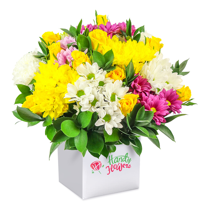 In this image, a dense, generously rounded bouquet sits neatly in a modern white square box, all placed against a completely white background that allows the colours to shine without distraction. The flowers create a harmonious blend of warm yellow, pure white, and bold fuchsia, all held together by a base of deep green foliage. Dominating the centre are large yellow chrysanthemums, with many fine petals radiating out like small suns, intertwined with sprays of yellow roses whose buds and partly opened blooms add intricate texture. Threaded through this golden heart are white daisy-style chrysanthemums with clear green centres, their clean, simple faces adding a fresh note, along with fuller white chrysanthemums that provide fluffy volume and a sense of softness. Fuchsia and magenta chrysanthemums are scattered throughout, some with subtle pink gradations, forming vivid streaks of colour that draw the eye around the arrangement, reminiscent of the vibrant displays seen in Highgate's independent shops and weekend markets. Hidden among the larger blooms, smaller pink and yellow rosebuds suggest unfolding beauty and ongoing freshness. The foliage is a deep, glossy green, likely sturdy leaves selected to cradle the flowers and fill the bouquet, ensuring it appears lush from every angle. The white cube box is both practical and stylish, acting as gift box and vase in one, and would sit comfortably on a café counter near Highgate Station or on a work desk overlooking the Heath. The overall impression is of an accessible, cheerful arrangement that brings a sense of celebration and colour without feeling overly formal or expensive.