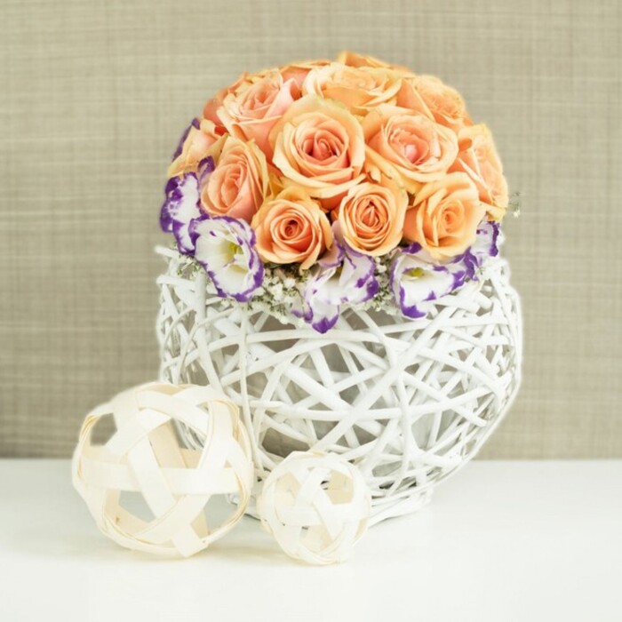On a pristine white tabletop, a beautifully balanced floral centerpiece takes centre stage, resembling something you might find at a sophisticated celebration along the streets of Highgate or near the quiet paths of Highgate Cemetery. Rising from the middle is a tightly composed dome of roses in dreamy shades of peach, apricot, and soft salmon-pink. Each rose is fully open, its velvety petals unfolding in spirals, and together they form a smooth, rounded mound that suggests abundance and harmony. Nestled snugly around the base of the roses is a ring of lisianthus flowers, bright white with ruffled petals edged in a rich purple hue, almost like ink tipped along each petal's edge. Delicate strands of white baby's breath are woven between them, bringing a faint, cloud-like texture that adds depth and lightness. The flowers are housed in a distinctive, bulbous white container, crafted from interwoven wicker or rattan strands that crisscross into an open lattice structure. This airy weaving lets light filter through and hints of stems show beneath, while the crisp white colour provides a modern, clean frame for the soft florals. Positioned on the white surface near the arrangement are two decorative spheres, one larger, one smaller, formed from thin, natural wood-toned strips. Their loosely woven, airy design echoes the texture of the main vessel and completes the composition with a relaxed, sculptural touch. Behind everything, a beige or oatmeal-coloured fabric backdrop, lightly textured like linen or fine burlap, gives a warm, neutral context. The overall feeling is one of calm romance and understated luxury, ideal for wedding tables, intimate engagements, or refined home décor for those in Highgate who enjoy natural materials and graceful, seasonal blooms.