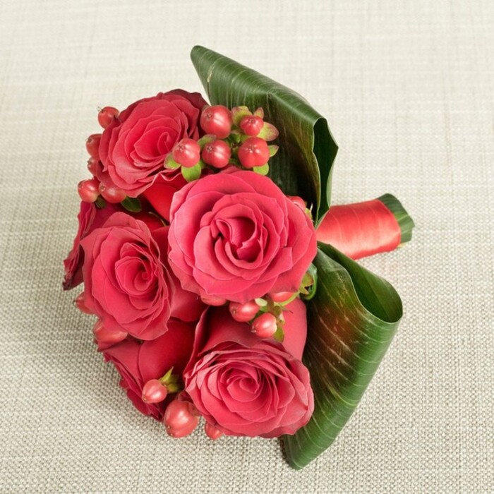 Set against a pale beige, delicately textured fabric backdrop that suggests a refined tablecloth or draped textile, a compact bouquet of rich red roses becomes the striking focal point of the scene. The arrangement, viewed from above at a slight angle, reveals around six roses clustered closely together, their velvety petals unfolding in intricate spirals that range from deep, wine-like crimson at the core to a vibrant scarlet along the outermost edges. Nestled intimately among these blooms are clusters of small, round berries, reminiscent of Hypericum, in hues of deep red and rosy pink, each berry smooth and glossy, catching the soft ambient light like tiny droplets. Embracing the flowers, several broad, dark green leaves are curved and folded with intention, their sturdy structure and defined veins lending both support and a sense of modern, sculpted elegance. Peeking from one side, a band of luxurious red satin ribbon encircles the stems, its gentle sheen echoing the richness of the petals and hinting at the skilled hand-tied craftsmanship beneath. The lighting is soft and even, as if filtered through a Highgate bay window on an overcast afternoon, casting minimal shadows and allowing colour and texture to stand out in harmonious balance. The mood is distinctly romantic and refined, suggesting a bouquet perfect for anniversaries, intimate dinners in a Highgate townhouse, or a polished bridesmaid posy carried along the leafy streets near Hampstead Heath. Every element-from the precise placement of berries to the graceful curve of each leaf-speaks of thoughtful design and an understanding of how classic red roses can be styled to feel both timeless and contemporary.