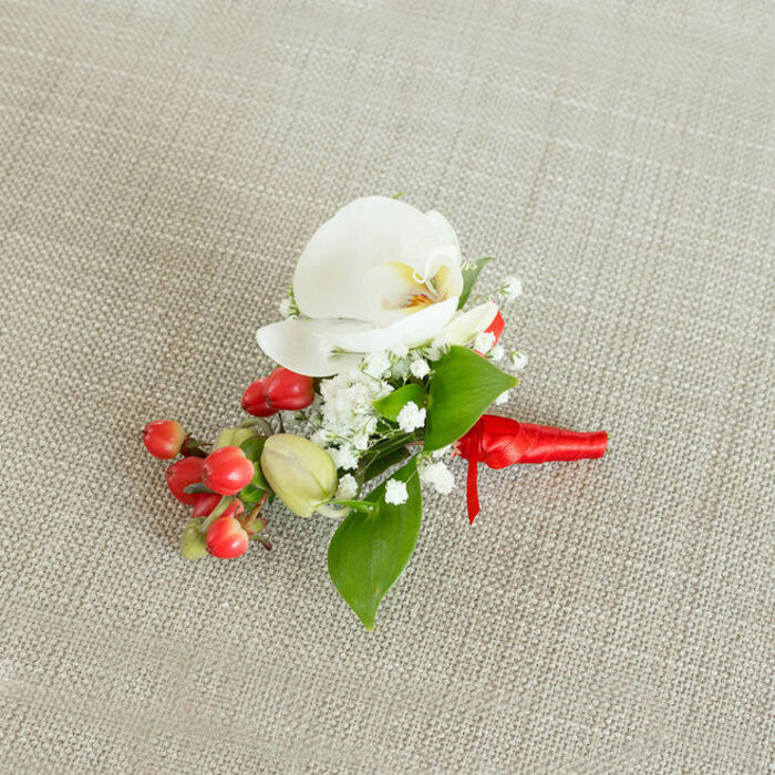 A single, exquisitely crafted floral boutonniere is photographed from above, resting on a pale, oatmeal-toned, woven textile that brings to mind the rustic elegance of a Highgate townhouse interior. At its center, a white Phalaenopsis orchid takes pride of place, its petals smooth and gently arched, forming a soft oval shape that feels both modern and timeless. Within the orchid's throat, a subtle blend of creamy yellow and muted orange glimmers, adding a warm focal point that contrasts gently with the cool purity of the surrounding petals. Embracing the orchid, clusters of fine white baby's breath appear almost weightless, their numerous tiny blossoms forming an airy halo that softens the overall silhouette. Just beneath this delicate veil of blooms, a tight grouping of glossy, cherry-red berries catches the light, introducing a bold pop of colour that suggests celebration - ideal for winter weddings or holiday events around Highgate and Crouch End. Interspersed with the red berries are a few smaller, pale green, oval buds, hinting at growth and new beginnings. Framing the entire composition, lush green foliage in slightly different shapes - some broader and more pointed, others long and narrow - adds depth and a fresh, garden-picked character reminiscent of local hedgerows near Queen's Wood. The stems are gathered cleanly and wrapped in a rich red satin ribbon, the material pulled smooth and secured with a compact, purposeful knot. Short ribbon tails extend just beyond the binding, creating a polished finish that clearly signals this piece is ready to be pinned on a lapel. The mood is elegant yet approachable, making this boutonniere perfect for grooms, groomsmen, or formal guests who want a refined floral touch crafted with the quiet sophistication associated with Highgate florists.