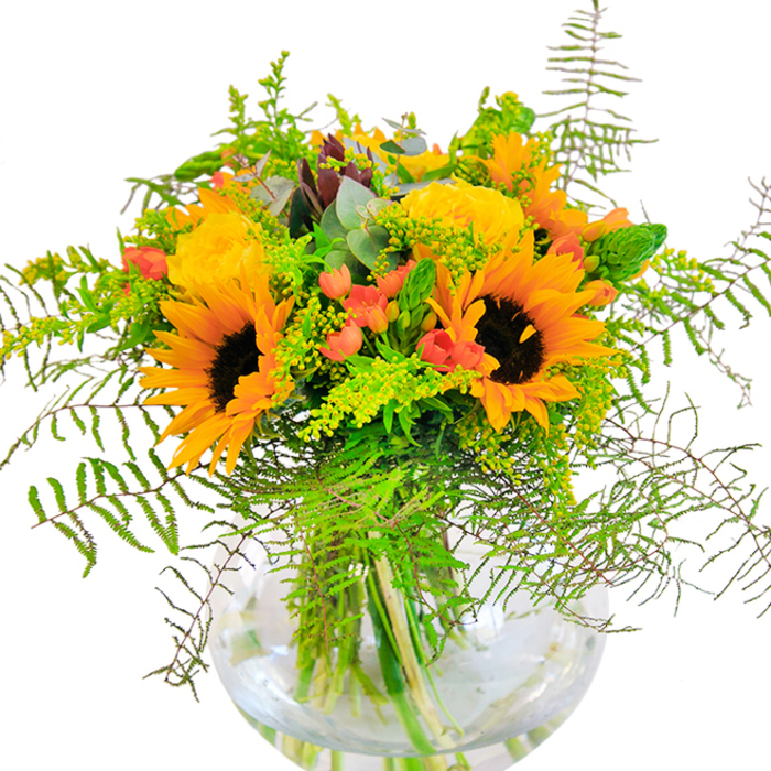 On first glance, this Highgate floral ensemble brings forth the vitality of sun-dappled afternoons by the ponds of Hampstead Heath. Towering sunflowers, ablaze with yellow-gold petals and dark, textured centers, command attention and speak of summer's peak. Clustered nearby, velvety yellow garden roses layer intricate spirals of petals, reminiscent of the elegant landscaping in Highgate's historic residences. Lush sprays of solidago and gentle spikes of filler emerge among the blooms, interspersed with the unexpected sparkle of red hypericum berries-vivid splashes echoing the lively markets along Highgate High Street. A tangle of ferns and trailing greenery cascades over the edge of the airy glass vase, infusing the whole with a sense of freshness and natural abundance. Cool eucalyptus fronds, striking in their subtle blue-green cast, weave through the arrangement, adding aromatic dimension and a hint of rustic sophistication. The gathered stems, visible in the clear water, evoke a summertime bouquet freshly picked from the edge of Queen's Wood. Flooded with natural daylight, each element glows, making this arrangement a superb gift for Highgate celebrations-whether marking an anniversary, welcoming a new neighbour, or simply bringing a thoughtful touch to a family gathering.