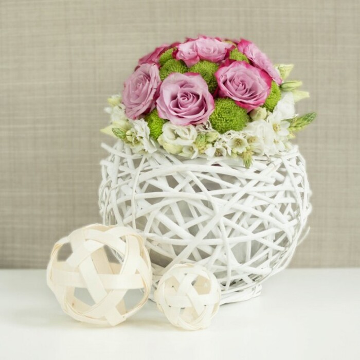 A thoughtfully styled centerpiece sits on a pristine white surface, looking as though it has been set down in a quiet Highgate home just before guests arrive. The focal point is a round, sculptural vase constructed from intertwined, twig-like strands painted white, their open, basket-style weaving allowing glimpses of shadow and air between each curve. This airy vessel cradles a compact floral dome that feels both abundant and tidy. At the heart of the bouquet are roses in soft pink and dusky lavender shades, many of them fully open so their layered petals form generous rosettes, with edges touched by slightly darker pink. Tucked between these pastel blooms are bright green button mums, whose small, cushion-like heads provide contrast in both shape and colour, bringing a fresh, lively accent that recalls the greenery edging Highgate's quiet side streets. Around the base and nestled amongst the roses and mums, fine white filler flowers weave through, creating a delicate froth that softens the arrangement's outline and gives it a subtle, cloud-like quality. To the left of the vase, two light-toned decorative spheres made from woven wicker or slim wooden strips rest casually on the same white surface, their open, crisscrossing patterns visually echoing the vase's design. Behind everything, the backdrop is a warm beige fabric, slightly textured and intentionally blurred so it serves as a neutral canvas for the flowers' colours and the intricate weaving of the decorative elements. The gentle, even lighting highlights the texture of each petal and strand, suggesting a calm, romantic mood that would suit a wedding reception in Highgate, a special anniversary dinner, or a refined brunch table dressed with fresh local flowers.