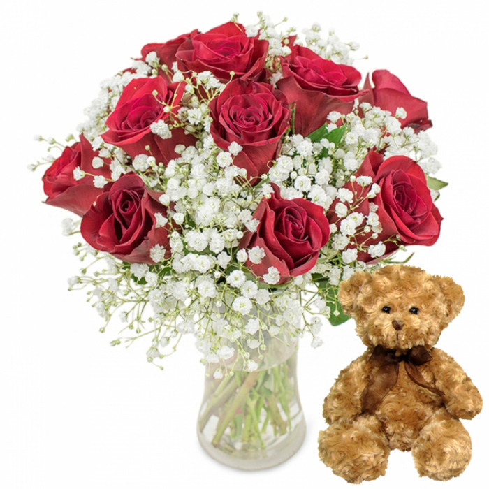 A beautifully balanced studio photograph features an arrangement that feels both luxurious and approachable, the sort of gift a Highgate resident might choose to mark an anniversary or romantic milestone. The left side of the frame is dominated by a clear glass vase containing a well-proportioned bouquet of about twelve red roses. The vase is straightforward and cylindrical, partially filled with fresh water, through which the green stems are visible, criss-crossing gently as they meet at the base. Above the vase's rim, the roses gather in a full, rounded crown, each flower displaying thick, velvety petals in a deep scarlet that verges on burgundy in the shadowed folds. Some blooms are open enough to reveal intricate layers, while others still cradle their centres in a more compact rosette, suggesting freshness and longevity. Threaded among the roses, fine clusters of white baby's breath introduce a soft, cloud-like texture, with countless tiny blossoms perched on delicate, branching stems. These white accents create a gentle halo effect around the roses, brightened further by a few glimpses of dark green leaves peering through the design, hinting at the bouquet's garden origins. On the right, a plush teddy bear sits upright, its light brown fur appearing slightly tousled and extremely soft to the touch. Rounded ears, padded paws, and a softly rounded belly give the bear an endearing, childlike silhouette, while its small, shiny eyes and carefully stitched nose and mouth create a sweet, calm expression. A slender dark brown ribbon is tied neatly at its neck, providing a simple yet elegant finishing detail. Set against a pure white background, the arrangement exudes clear, focused emotion: romance, warmth, and gentle nostalgia, perfectly suited to imagining a surprise flower delivery to a Highgate home overlooking Waterlow Park or a shared celebration after a walk up to Parliament Hill.