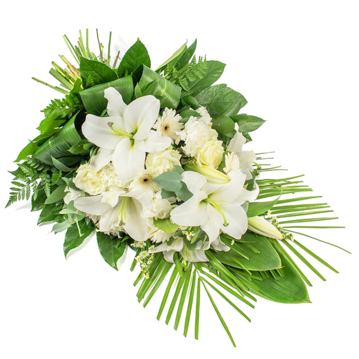 This photograph captures a dignified white and green funeral spray arranged on a strong diagonal, as if ready to rest alongside a coffin or at the front of a memorial space in Highgate. The composition has a clear front-facing design, densely packed with blooms in the centre and carefully layered foliage framing the edges. Dominating the arrangement are several large white lilies, their trumpet-shaped flowers open wide and facing slightly outward, each petal satiny and gently curved, giving the impression of softness and light. In between these prominent lilies stand firm, elongated buds, their pale green casings hinting at the blossoms still to emerge. Around the lilies, clusters of white roses in varying stages of bloom contribute a timeless, elegant feel; some buds are tightly wrapped, while others reveal loosely overlapping petals that form soft cups of colourless light. Ruffled white carnations bring a reassuring fullness, their many-edged petals creating small, cloud-like mounds that nestle comfortably around the roses and lilies. Scattered among them are smaller, daisy-like white flowers, likely gerberas or similar varieties, with neat rings of petals surrounding subtle cream or pale lemon centres, breaking up the larger forms and adding gentle contrast. Fine white filler flowers are woven throughout, giving a filigree delicacy to the overall design. The flowers rest on a base of mixed greenery: broad, glossy dark green leaves arranged in layers, light feathery ferns that soften the profile, and distinctive round eucalyptus leaves shaded in muted blue-green, lending a contemporary undertone. At the upper section, long, blade-like leaves are tightly wrapped and secured, forming a tidy upright feature. From the lower right corner, a fan of slender palm fronds sweeps outward and downward, extending the composition into a graceful point. The spray radiates peace, respect, and remembrance, a fitting floral expression for a farewell in the Highgate area, perhaps delivered to a quiet ceremony near Highgate Hill or the pathways of Waterlow Park.