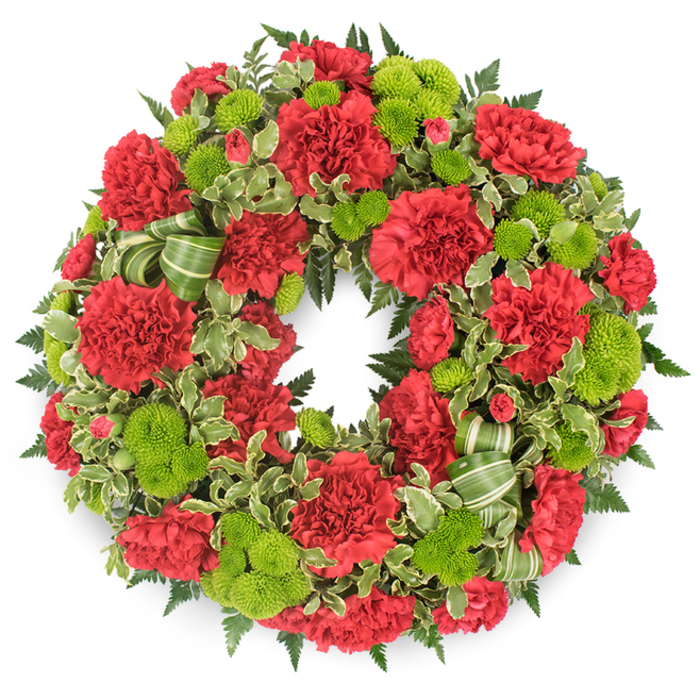 A striking circular wreath for a funeral or memorial is photographed against a clean, white background, highlighting every detail of its red and green composition as if it were thoughtfully placed on a stand in a Highgate church. The wreath's ring is fully cloaked in flowers, with luxurious red carnations forming the main body. Their densely layered, frilled petals create a soft yet sculptural surface, each bloom slightly different in size and openness, suggesting the unique stories that gather around a life. Between the carnations, vivid lime green button chrysanthemums are dotted evenly throughout the circle, their perfectly rounded, compact heads made of countless tiny florets that lend structure and brightness. The play of crimson and chartreuse creates a dynamic but respectful palette, strong enough to stand out in a chapel yet gentle enough to convey tenderness and solace. Encircling and threading through the flowers, an assortment of lush foliage establishes depth: dark, fern-like fronds project just beyond the blooms, weaving a subtle shadowed frame, while variegated pittosporum leaves with creamy white edges introduce lighter, almost painterly strokes through the greenery. In the lower quadrant, a graceful bow or ribbon feature in alternating shades of light and dark green loops outward, blending with the leaves and echoing the natural hues of Highgate Wood and the slopes around Parliament Hill. The lighting is even and tranquil, throwing a delicate shadow beneath the wreath that gently lifts it from the background and accentuates its circular form. The entire design radiates a quiet dignity and caring craftsmanship, a fitting floral expression of sympathy and remembrance for services, wakes, and graveside tributes across the Highgate community.