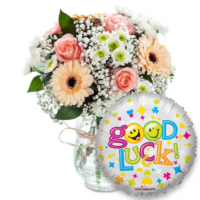 A locally minded good-luck arrangement, composed with the kind of care regulars expect from a Highgate florist. The bouquet, set in a clear glass vase finished with a rustic twine bow, blends pale pink roses whose petals feel satiny and close-set with cheerful peachy-orange Gerbera daisies that have rich brown centres and a slightly velour texture. Interspersed among them are fluffy sprays of white baby's breath (Gypsophila) that create a soft, lace-like cloud, while compact white and lime-green chrysanthemum florets add a bright, lively punctuation. Dark green foliage provides contrast and depth so the pastel hues remain luminous under soft lighting. In the right foreground a round metallic silver Mylar balloon overlaps the vase; it's patterned with scattered colourful stars, hearts and confetti, and carries the exuberant message Good Luck! in playful multi-coloured letters. The word Good shows the two o's replaced by animated yellow smiley faces-one winking, the other tongue-out-adding a lighthearted, encouraging character, while Luck! continues in a sequence of vibrant greens, oranges, reds and blues. A discreet brand mark sits near the balloon's base, and the whole scene is photographed on a white ground to accentuate the fresh colours. This arrangement reads as a hopeful, celebratory gesture suited to farewells, new jobs or exam days across Highgate Village, around Highgate Hill and toward Hampstead Heath; it's the sort of composition Flowers Highgate might craft for neighbours offering a bright, reassuring send-off.