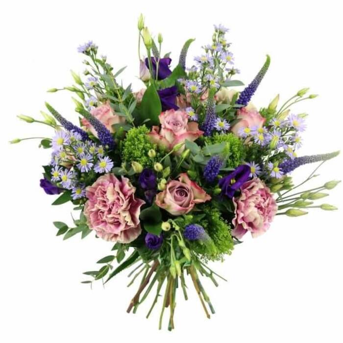 The image presents a meticulously arranged, dome-shaped bouquet of fresh flowers placed against a spotless white background, showcasing every shade and texture with clarity. Dominating the arrangement are soft pink and mauve roses, some still in bud with outer petals wrapped protectively around the core, others more open so that the layered spirals at their centres are clearly visible. Their petals shift delicately in tone from deeper pink at the heart to paler, almost cream edges, creating a gentle ombré effect. Interwoven with the roses are large, ruffled carnations in dusty rose and purple-mauve hues, their densely packed, frilled petals adding a sense of fullness and luxurious texture. Rising from within the blooms are tall, slender spikes of deep lavender-blue Veronica, which lend vertical interest and a touch of wild garden charm. Around them, smaller clusters of light purple, periwinkle-toned aster-like flowers with tiny yellow centres form a soft, cloud-like layer throughout the bouquet, lightening and balancing the richer tones. Nestled closer in are several darker purple lisianthus blossoms, their smooth, cup-shaped petals providing contrast in both shape and depth of colour. The whole design is anchored by an abundance of foliage, including broad, silvery-green eucalyptus leaves that arc gently outward and distinctive tufts of bright, chartreuse-coloured green filler. These vibrant greens punctuate the arrangement with lively bursts of colour and add a fresh, almost dewy feeling. The stems beneath are clean, straight, and tightly bound, confirming the hand-tied style, ready to drop into a favourite vase in a Highgate sitting room or hallway. The composition speaks of skilled craftsmanship and quiet romance, ideal for luxury flower delivery, milestone birthdays, or anniversary celebrations around Highgate Village, Dartmouth Park, and the leafy streets leading towards Parliament Hill.