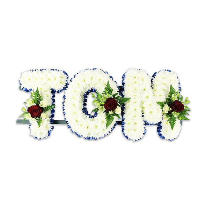 The word TOM is rendered here as a dignified, three-dimensional floral tribute, each letter meticulously covered in countless small white chrysanthemums so that the surface reads as a continuous, cushiony expanse of petals. Set against a neutral white background, the letters are clean and sculptural; the chrysanthemums' tiny textures catch light in soft highlights and shadows, lending a tactile calm. A striking royal blue ribbon outlines every edge with a steady, even line that gives the composition a formal clarity and a cool contrast to the warm whites.

Each letter is accented by a composed spray placed toward the centre or lower portion: a solitary, deep red rose serves as the focal heart of each cluster, its velvety bloom symbolising love and remembrance, while delicate creamy-white spray roses and arcing fern fronds provide contrast and graceful movement. The arrangement's green foliage-slender ferns and glossy leaves-adds depth and keeps the overall silhouette serene and balanced. A dark green or black metal bar is visible beneath the base, the subtle engineering that ensures the letters stand upright for display. This piece is the sort of commemorative tribute often seen at services for families from Highgate Village and the surrounding streets by the Pond, offered with the careful finishing touches a local florist would apply to honour a life.