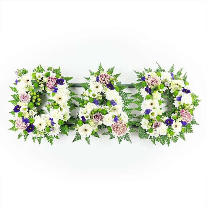 This photograph presents an expertly designed floral tribute spelling out DAD in three large, block-style letters, each one constructed from a profusion of white and purple funeral flowers. The letters are aligned horizontally and set against a minimalist white background, giving the arrangement a calm, uncluttered feel. At the heart of each letter lies a thick cushion of white chrysanthemums and daisy-like flowers, their tight heads pressed closely together so that the mechanics beneath are fully concealed. The whites vary subtly in tone, giving the surface a lively yet harmonious appearance, as if catching soft daylight similar to that filtering through the trees in nearby Highgate Wood. Among this white base, the florist has thoughtfully placed small sprays of lavender and mauve flowers-roses, carnations, and perhaps lisianthus-whose delicate petals add a gentle, romantic note. Deeper purple blossoms, resembling violas or miniature pansies, bring depth and a hint of regal solemnity, reminiscent of the rich hues often seen in the stained glass of Highgate's churches. Surrounding each letter is a collar of lush green foliage, including feathery ferns and firm, glossy leaves, which frames the shapes and provides structure so that the D, A, and D are instantly legible. The green has a fresh, woodland character, echoing the natural beauty of Waterlow Park and the slopes leading up to Highgate Village. The overall lighting is bright but soft, with no harsh glare, allowing viewers to appreciate the meticulous placement of each stem and the overall balance of colour and texture. This DAD floral lettering is a classic, meaningful tribute, particularly suited to funerals and memorials in and around Highgate where families wish to present a clear and affectionate final message to a beloved father.