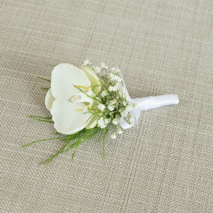 A refined white orchid boutonniere is shown resting on a neutral, light beige background, the fabric's visible weave adding a gentle rustic texture that contrasts beautifully with the polished elegance of the flowers. At the centre sits a flawless Phalaenopsis orchid bloom, its generous, overlapping petals a cool, luminous white that appears almost sculpted. The petals open with a natural, relaxed curve, revealing a soft hint of pale yellow and cream in the orchid's throat, where the central column and tiny markings create delicate, intricate interest. Surrounding this focal bloom is an airy halo of white baby's breath, each tiny clustered flower forming a cloud of softness that lightens the overall silhouette of the boutonniere. Fine, wispy strands of vibrant green foliage peek out between the baby's breath, their feather-light leaves adding a fresh, botanical touch that hints at early morning walks through Highgate's leafy side streets and gardens. The stems of the orchid, gypsophila, and greenery are carefully aligned and bound together with a band of pristine white satin ribbon, wrapped smoothly and evenly, then finished with a modest, elegant bow that sits neatly at the base. The ribbon's subtle sheen catches the soft light, echoing the gentle gloss of the orchid petals. The lighting in the image is calm and even, casting delicate shadows that give depth to the arrangement without harsh contrast, allowing the viewer to sense both the silkiness of the petals and the fine, crisp texture of the fabric beneath. The overall impression is of classic, minimalist sophistication: a boutonniere ideal for a groom or groomsman attending a wedding at a local Highgate venue, blending contemporary style with timeless romance, and evoking the quiet charm of this North London neighbourhood. Crafted in this way, it feels like the kind of piece a careful florist would prepare for same-day delivery to a townhouse overlooking the tree-lined streets near Highgate Tube.