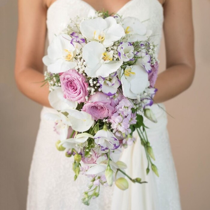 Against a blurred canvas of warm beige, evocative of the golden hour over Highgate Ponds, this photograph captures a bride-visible from her delicate, lacy gown's bodice downward-tenderly holding an opulent, teardrop-shaped bridal bouquet. Every flower has been thoughtfully chosen by Flowers Highgate to reflect the heritage and refined taste of the North London community. Majestic white phalaenopsis orchids crown the bouquet, their petals generous and creamy, each centre tinged with sunshine yellow that brings the display to life. Plush, romantic garden roses in muted pink unfurl just below, interlocking with clusters of lavender and lilac-hued lisianthus with ruffled edges, conjuring images of meadows along Hampstead Lane. Wisps of white baby's breath, like a sudden mist over Waterlow Park, fill any gaps and create a gentle, floating effect, enhanced by minimal but deliberate greenery that outlines the bouquet's elegant flow. The arrangement cascades purposefully, inviting the eye to follow its path down towards the bride's gown, echoing the natural slopes and woodland paths of Highgate. This bouquet radiates tranquility and modern grace, holding within its design an undercurrent of old-world romance-making it an exquisite choice for wedding ceremonies inspired by Highgate's storied landscapes and artistic sensibility.