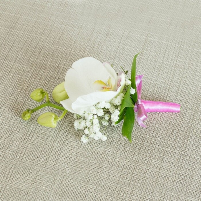 An exquisitely crafted floral boutonniere is displayed horizontally on a pale beige, linen-like fabric, the slightly nubby texture of the material lending a quiet, natural elegance that recalls the soft interiors of homes around Highgate Village. The composition is centred on a single, immaculate white Phalaenopsis orchid, its generous petals opening outward in a gentle curve. The flower's centre glows with subtle yellow notes and the faintest whisper of blush pink, drawing attention to its refined, sculptural beauty. Nestling closely at the base of the orchid, tiny sprays of white baby's breath form a delicate, clouded surround. Each minute bloom adds airiness and volume without overwhelming the main flower, giving the whole piece a dreamy, romantic quality reminiscent of early morning light over nearby Hampstead Heath. Slim, bright green leaves slip out from behind the central cluster, their smooth, linear form and vivid hue offering a fresh counterpoint to the soft whites. To one side, two or three lime-green orchid buds remain tightly closed along a slender stem, suggesting growth, continuity, and the unfolding of new memories on a wedding day or prom night. The stems are neatly gathered and wrapped in a narrow band of lustrous fuchsia satin ribbon, tied into a small yet perfectly proportioned bow. The ribbon catches the light with a gentle sheen, its bold pink colour injecting a joyful, celebratory note into the otherwise calm palette. Soft, even lighting enhances every texture-the weave of the fabric, the satin's gloss, and the orchid's velvety petals-creating an image that feels intimate, handcrafted, and ideal for describing a groom's boutonniere or corsage styled for a special Highgate event with timeless, polished grace.