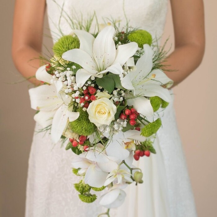 Set against the refined lace of a wedding dress and the distinct, peaceful foliage of Highgate, this elaborate cascading bridal bouquet emanates both grandeur and gentle intimacy. The bouquet's principal stars are striking white lilies, their trumpet-shaped blooms expansive and lightly freckled, reminiscent of the classic blooms favored for elegant weddings at Kenwood House. At the very centre, a creamy white rose unfurls, a symbol of enduring affection. Interspersed throughout the arrangement, brilliant lime-green cushion chrysanthemum blooms add flashes of cheerful colour, much like the lively market stalls in Highgate Village on a sunny day. The tender spray of white baby's breath softens every angle, emerging like pockets of morning fog over Hampstead Heath. Accents of vibrant red Hypericum berries are strategically nestled among the florals, providing a fresh and festive note recalling the area's seasonal hawthorn berries. The bouquet's cascading silhouette is gracefully defined by streams of white Phalaenopsis orchids, whose delicate forms evoke the serenity of Highgate's secret woodlands. Finished with slender green fronds and glossy leaves, the arrangement's artistry is brought together with textural depth and a sophisticated sense of motion. The figure of the bride gently clutching the handle grounds this exuberant floral celebration in real wedding tradition, making it a perfect choice for those drawn to Highgate's unique blend of historical charm and natural beauty.
