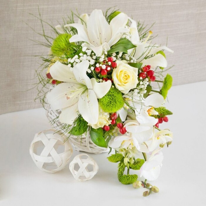 Set against a softly textured beige backdrop, an artfully designed floral arrangement in a white woven basket appears ready to grace a Highgate dining table or wedding reception at a local venue. The upper portion of the composition is dominated by several large white lilies, their elongated petals curving outward to display subtle speckling and soft golden stamens, conveying a sense of light and openness. Tucked among these lilies, a single pale yellow rose in full bloom provides a gentle focal glow, its silky petals layered in a perfectly formed spiral that suggests tenderness and romance. From the right side of the basket, a flowing curtain of white phalaenopsis orchids cascades elegantly downward, the broad, smooth petals of each bloom giving the impression of delicate porcelain, forming a graceful teardrop shape. Surrounding the main flowers is a lush mix of foliage-fine, airy greens, slender grasses, and slightly broader leaves-that build depth, with some stems arching outward as though caught in a soft Highgate breeze. Bright green pom-pom chrysanthemums and moss-like dianthus balls add texture and cheerful colour, popping against the predominantly white palette. Dotted throughout are clusters of deep red berries, rich and glossy, adding a festive warmth that would suit winter celebrations near Hampstead Heath or around the leafy Highgate streets. The arrangement sits on a clean white surface that subtly reflects the colours above, enhancing the feeling of freshness. To the left, two decorative white spheres woven from slender strands echo the basket's texture and introduce a sculptural element. Overall, the composition conveys quiet sophistication, meticulous craftsmanship, and the sort of natural elegance one might expect from Flowers Highgate when creating a standout wedding table arrangement or special occasion centrepiece.