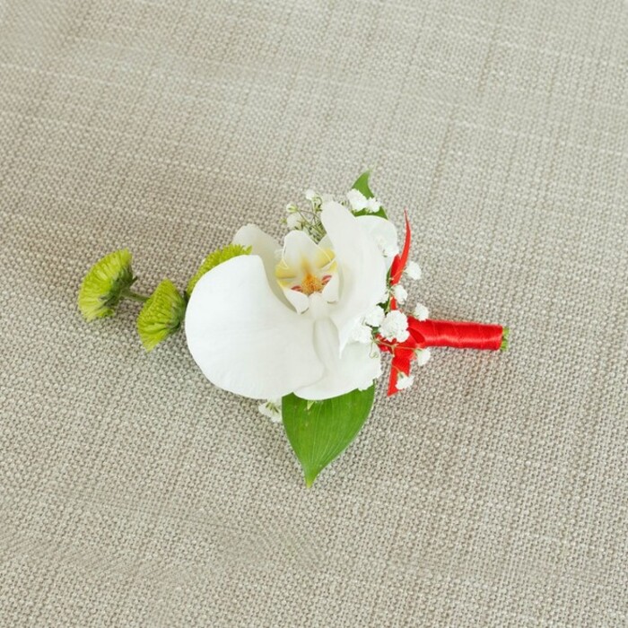 An artfully composed boutonniere is photographed close-up against a softly textured, light beige linen background, the fabric's gentle weave offering a calm, neutral canvas reminiscent of table linens in a Highgate village restaurant ready for a wedding breakfast. Dominating the composition is a large white phalaenopsis orchid bloom, its petals broad, smooth, and gently arching, with a faint sheen that suggests velvety softness under the fingertips. The orchid's centre is alive with detail: a soft yellow hue radiates from the throat, blending into fine touches of reddish-orange or amber, creating a subtle pattern that feels both natural and intricate. Framing this focal blossom, sprays of white baby's breath extend outward in airy clusters, their numerous tiny flowers catching the light like scattered stars, bringing a delicate, romantic haze to the design. To the left side, two distinct lime-green chrysanthemum buds-round, tightly packed, and almost moss-like-introduce an invigorating pop of colour and an intriguing, slightly spiky texture that contrasts the orchid's fluid lines. These buds mirror the fresh greens you might notice strolling past the gardens off Highgate West Hill in early summer. Beneath the flowers, a generous deep-green leaf provides a solid base, its surface smooth and slightly glossy, while a smaller, darker leaf nestles near the stems, subtly anchoring the composition. The stems are carefully bound in a vivid crimson red satin ribbon that hugs them snugly, its sheen reflecting soft highlights and culminating in a small, neatly arranged bow or knot. This red accent adds a bold yet tasteful note of celebration, ideal for a groom or groomsman at a formal ceremony. The overall mood of the image is refined and contemporary, with balanced colour, considered textures, and gentle, even lighting that highlights the craftsmanship and thoughtful design behind this special-occasion floral accessory.