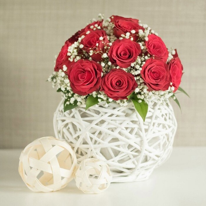 An elegant, contemporary floral composition is shown, ideal as a wedding table centrepiece for a Highgate venue, with a tight bouquet of red roses arranged in a sculptural white vase. The roses are a deep, classic crimson, their velvety petals unfolding in perfect spirals, each bloom open and full but still crisp at the edges, suggesting they have been freshly arranged. Gathered closely together, the flowers create a smooth, dome-shaped cluster, so that the viewer's eye reads them almost as a single, luxurious cushion of red. Woven between them, airy sprays of white baby's breath introduce a light, cloudlike texture, brightening the arrangement and echoing the delicacy of bridal fabrics and soft candlelight. A few small, dark green leaves nestle at the base, barely visible yet important in lending a natural balance to the palette. Cradling this floral dome is a bulbous, bright white vase, fashioned from interlaced strips that form an open, basket-style weave, reminiscent of artisanal pieces that might be found in a carefully curated Highgate interior. The vase is placed squarely on a smooth, pale surface, perhaps a linen-covered table in a restaurant above Highgate Hill, ready for guests to sit down to dinner. Just to the left of the vase, two decorative orbs crafted from flat, off-white woven bands are positioned with intention: the larger sphere nearer the front, the smaller partially hidden behind, adding dimension and reinforcing the tactile, woven theme. Behind everything, a softly textured neutral wall, slightly out of focus, creates a gentle backdrop that allows the rich red roses and crisp white elements to command attention. The overall mood is serene and romantic, perfect for intimate weddings, engagement parties, or anniversary celebrations across Highgate and nearby Crouch End.