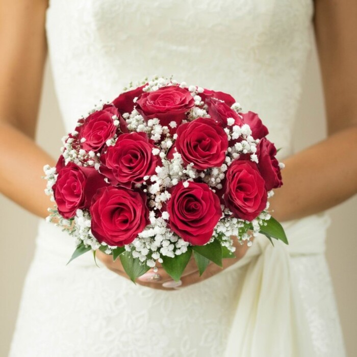 Set against the stately charm of Highgate's leafy avenues, this close-up image captures the essence of wedding day elegance. At mid-torso, a bride in a lavishly embroidered white gown embraces a rounded bouquet overflowing with deep red roses, each blossom unfurling in textured layers of velvet. These passionate flowers are punctuated with clouds of crisp white gypsophila, adding both sophistication and an ethereal softness-like morning mist in Highgate Wood. A few luminous green leaves peek from beneath, grounding the bouquet in nature's freshness. The bride's graceful hands with manicured nails secure the stems just so, while the dappled fabric of her gown-a weave of lace and satin-reflects the light, amplifying the bouquet's romantic drama. All fades to a creamy blur in the background, directing the eye to every floral detail and the serene confidence of bridal preparation. Crafted in the heart of Highgate by specialists attuned to the area's unique style, this bouquet is ideally suited for articles investigating wedding flowers, icons of romance, or the craftsmanship behind N6's most memorable celebrations.