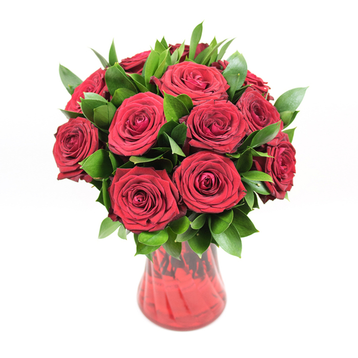 In this image, a beautifully composed bouquet of deep crimson roses takes centre stage, displayed in a translucent red glass vase against a flawless white backdrop that lets every detail stand out. Around twelve to fifteen roses are arranged in a rounded, compact cluster, each bloom fully open yet still firm, revealing concentric rings of velvety petals that spiral inward to a rich, shadowed heart. The red is intense and saturated, more like a polished garnet than a bright scarlet, with subtle shifts in tone where the petals overlap and the light softens towards the edges. Nestled throughout the arrangement is a generous collar of dark, glossy foliage-likely ruscus or similar greenery-its smooth, pointed leaves adding texture and a deep green contrast that makes the red appear even more vivid. The foliage emerges between the flowers in layers, giving the bouquet fullness and a well-balanced, almost sculpted appearance, like something you might see thoughtfully styled in a Highgate village window. The vase itself is an integral part of the design: a rich, red glass container with a gently curving silhouette that narrows at the middle and slightly flares at the rim. Its surface has a subtle ribbed or swirled pattern, catching the light and creating ripples of darker and lighter red tones, echoing the movement within the petals above. The stems are only just visible through the tinted glass, adding a hint of natural structure without drawing attention away from the flowers. With the pure white studio background keeping the focus sharply on the interplay of colour and texture, the whole arrangement radiates warmth, romance, and quiet luxury-an ideal visual for celebrating anniversaries, Valentine's surprises, or meaningful gestures of love delivered across Highgate and the surrounding streets. Crafted with the eye of an experienced florist, it speaks of care, intention, and enduring affection.
