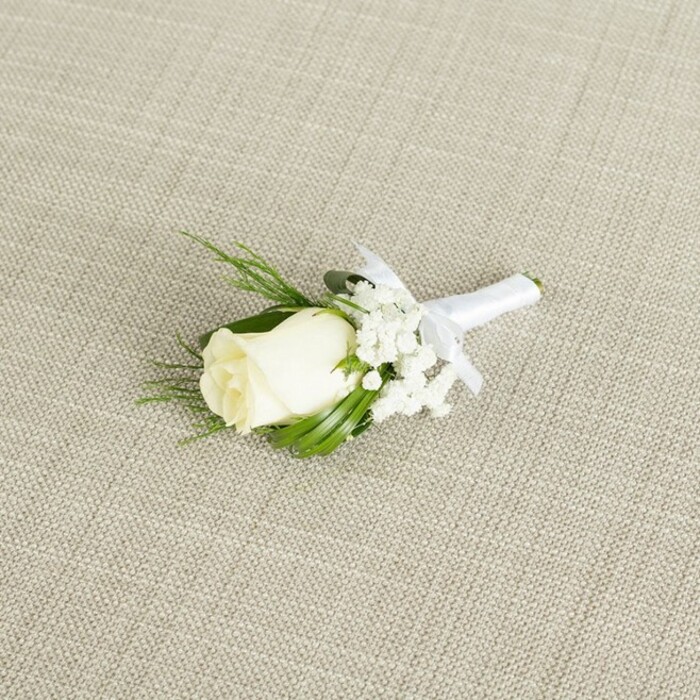 A refined white rose boutonniere is displayed at a gentle diagonal across a pale beige, linen-like fabric, its woven texture forming a discreet, natural backdrop that lets the florals take center stage, much like a well-set table at a Highgate celebration. The focal point is a single, tightly furled rosebud in soft ivory-white, the petals smooth, velvety, and closely wrapped, with faint green tinges at the base where the sepals cradle the bloom, hinting at its freshness. Surrounding the rose are light, cloud-like clusters of white baby's breath, their tiny, star-shaped flowers fanning out delicately and creating a soft halo that enhances the rose without overwhelming it. Interspersed behind and beside the blooms are slender, pointed green leaves and a couple of feathery, fern-like sprigs, the foliage vibrant and rich in tone, adding depth and a garden-fresh character that calls to mind early-morning walks through Highgate Wood. The stems of the rose and supporting flowers are neatly aligned and wrapped with immaculate white satin ribbon, the fabric smoothly and tightly wound to form a polished handle. A small, symmetrical bow made from the same satin sits just beneath the head of the rose, its tidy loops and short tails suggesting careful, practiced craftsmanship. The light in the image is soft and diffused, revealing subtle shadows along the folds of the petals and a gentle sheen on the ribbon, without any glare or harsh contrast. The overall effect is one of classic, timeless elegance-an understated yet meaningful accessory that would suit a groom or groomsman at a wedding ceremony held in one of Highgate's historic churches or intimate local venues, capturing the purity, new beginnings, and quiet joy of the day in a single, beautifully composed floral piece.