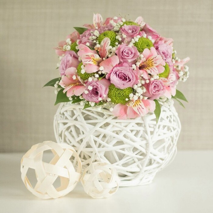 This image captures a beautifully styled floral arrangement that feels at once contemporary and romantic, as if prepared by an experienced Highgate florist for a light-filled sitting room near Waterlow Park. Centered on a spotless white surface, the bouquet rises from a distinctive white vase made of slender, overlapping strips that curve around each other to form an open, basket-like lattice. Its rounded, slightly bulbous silhouette provides a sturdy yet airy base, allowing glimpses of stems within and adding a sculptural touch to the composition. Overflowing from the top is a lavish mix of flowers in a calm yet joyful palette of pastel pink, soft lavender, bright lime green, and crisp white. Full, velvety roses in pink and lavender form the core of the arrangement, their petals layered like delicate fabric folds, suggesting fragrance and softness. Intertwined with them are elegant pink alstroemeria, their trumpet-shaped blossoms marked by fine stripes and speckles that draw the eye deeper into the bouquet. Small, round lime green button chrysanthemums punctuate the design with fresh, almost citrusy colour, creating a lively contrast against the more muted tones, much like a patch of new spring growth in Highgate Wood. Fine sprays of white baby's breath are scattered throughout, adding a gossamer lightness and filling the spaces between the larger blooms. Dark green leaves appear at intervals, framing and supporting the flowers without overwhelming the pastel serenity. To the left of the vase, two decorative woven spheres in natural, pale wicker shades sit on the same white surface, one larger and one smaller, echoing the latticework of the vase and adding visual rhythm. The softly textured beige wall behind keeps the focus firmly on the flowers and their handcrafted container, creating an atmosphere of elegant calm, suitable for wedding décor inspiration, refined home styling, or a thoughtful flower delivery for a special occasion in Highgate.