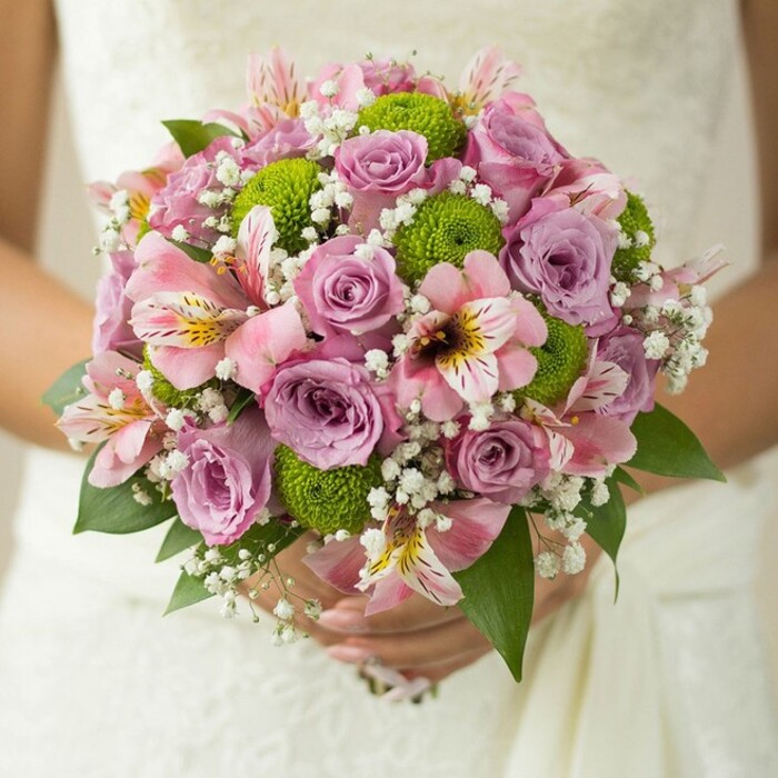In this beautifully intimate moment, a bride from Highgate softly holds a spherical bouquet, its floral artistry displayed against the subtle luxury of white lace fabric. Dominating the composition are roses in hues of soft blush, pale pink, and lavender, their delicate petals unfurling gracefully, some still dusted with tiny droplets, as though freshly gathered near Highgate's village gardens. The bouquet's trendy spirit is captured by punchy lime green button chrysanthemums, their petite, pom-pom shapes standing in cheerful counterpoint to the classic roses. Intertwined among these central blooms lie pink alstroemeria, trumpet-shaped and accented with dark pink and maroon striations, offering a gentle sense of the exotic. Wispy trails of baby's breath escape between the flowers, creating an airy volume and a halo-like effect that enhances the bouquet's lightness. Dark, glossy leaves peek from the base and sides, perfectly framing the composition. Seen through the eyes of a local artisan, this bouquet effortlessly fuses traditional wedding romance with the fresh, creative energy that defines Highgate. An ideal choice for ceremonies at St. Joseph's or evening receptions overlooking the city, it stands as a shining example of how local floristry brings personal dreams to life.