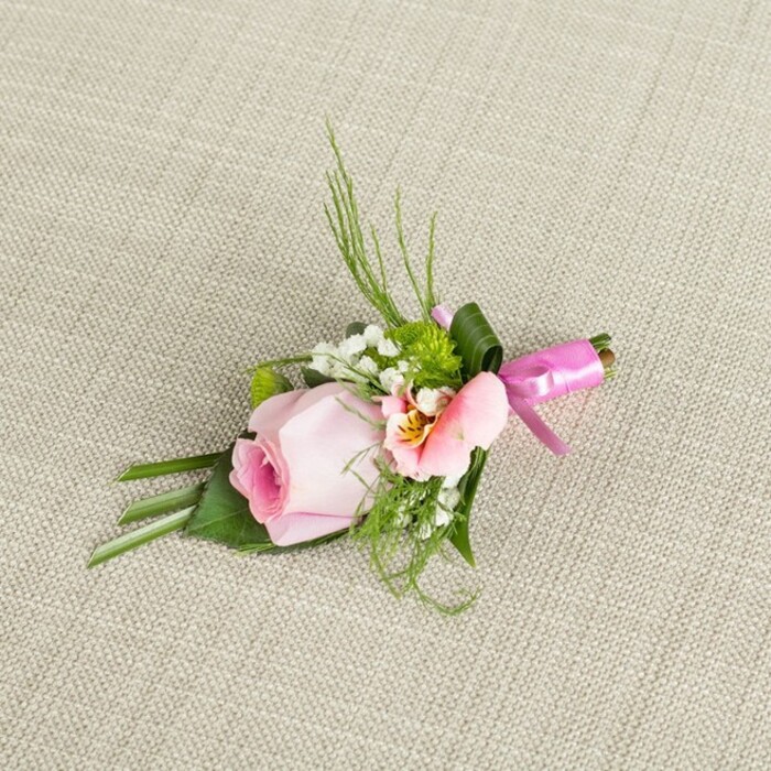Laid upon a textured, neutral linen reminiscent of Highgate's timeless architecture, this boutonniere tells a story of exquisite detailing and local craftsmanship. The arrangement centres on a softly blushing pink rose, spiralled tightly in the centre, its petals transitioning from roseate depths to pale outer edges that evoke first light over Hampstead Heath. Alongside, a lighter peach or pink ruffled flower-perhaps a fresh Alstroemeria-unfolds its petals, displaying subtle yellow nuances and fine dark markings that add a painterly touch. Miniature buds of baby's breath gather in gentle white accents, paired with vivid green globes of mini cushion mums which inject a contemporary twist and vivid textural interest. An array of foliage, from fine, upright grasses to supportive, dark broad leaves, imparts movement and substance, reminiscent of strolls through Highgate Wood or the lush gardens on Bisham Gardens. At the boutonniere's base, a glossy fuchsia pink satin ribbon ties all together, forming a small, precise bow whose celebratory accent nods to festive events on Highgate's bustling High Street. The clean, off-white woven background provides a classic foil, illuminating the arrangement's hues and the freshness of just-picked blooms. Whether destined for a groom's lapel, a prom, or a landmark event, this locally made floral accessory celebrates love, tradition, and the distinguished artistry of Highgate's florists.