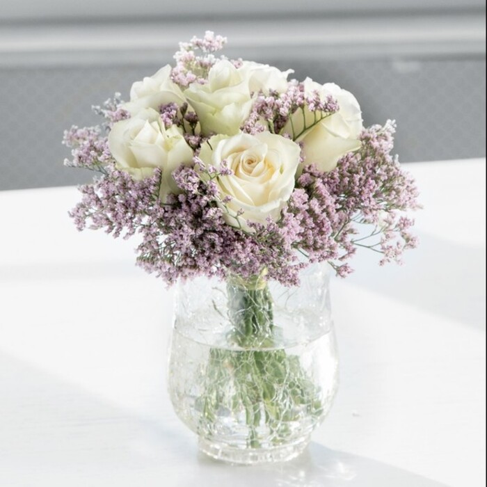 A beautifully composed bouquet of creamy white roses and lilac-tinted statice sits in the centre of the frame, exuding an air of quiet sophistication that feels at home in a Highgate townhouse or a light-filled flat near Pond Square. The arrangement is full and rounded, with approximately a dozen white roses forming a domed heart, their petals soft and velvety, opening gradually in elegant spirals. Each bloom carries an ivory-cream hue, subtly warmed by the daylight that filters gently across the scene, giving the flowers a soft inner glow. Weaving between the roses are plentiful sprays of lavender statice, their many tiny flowers forming a gossamer veil that curls and twists among the larger heads. This interplay of textures-the plush, almost buttery rose petals against the fine, papery statice-creates a rich visual and tactile contrast, suggesting both delicacy and structure. The stems are gathered tidily into a tall, clear cylindrical glass vase, where straight green stems are visible through the clean water, hinting at the freshness of a bouquet arranged only hours before. The vase rests on a sleek white surface that reflects subtle shadows and highlights, while the background is softly blurred, perhaps an interior wall or window, making the flowers the undisputed focal point. The overall mood is one of peaceful elegance, ideal for marking anniversaries, welcoming a new home, or offering a gentle note of sympathy. From the perspective of a florist, the proportions and balance speak of attentive craftsmanship and a desire to let classic flowers shine with minimal distraction, allowing their natural beauty and quiet fragrance to carry the emotion of the occasion.