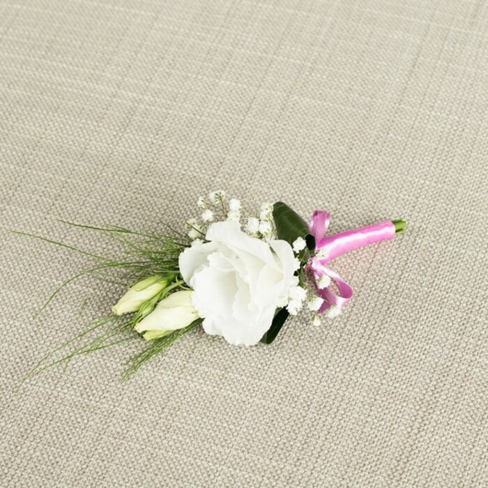 A refined white lisianthus boutonnière is photographed from above, resting diagonally on a light beige textile whose subtle weave suggests natural linen or cotton, its neutral tones reminiscent of calm interiors along Highgate's tree-lined streets. The main flower is a fully opened lisianthus, its petals a pure, luminous white, ruffled and layered so they fold into one another like soft waves. Positioned slightly off-centre, the bloom feels both delicate and confident, commanding attention without any harshness. Just below and to the left, two lisianthus buds extend gracefully, tightly furled with creamy ivory and pale green hints, capturing that poised moment right before they unfold. Interwoven among the blooms are delicate tufts of white gypsophila, each tiny flower adding a misty, cloud-like softness, as if echoing spring blossom drifting over Highgate Wood. The arrangement is framed by fresh greenery: one broader dark green leaf cradles the main flower from behind, while several slender, grass-like leaves sweep outward towards the left edge of the frame, lending an organic, slightly whimsical movement. At the base, the stems are carefully gathered and finished with a bright fuchsia satin ribbon, tied into a small but confident bow. The colour is vivid and joyful, a playful accent that immediately draws the eye and contrasts beautifully with the white petals and muted background. The soft, even lighting allows you to see every petal edge and fibre in the fabric, giving a sense of craftsmanship and care, perfect for a groom's lapel at an intimate Highgate wedding or a formal event that calls for understated elegance with a touch of personality.