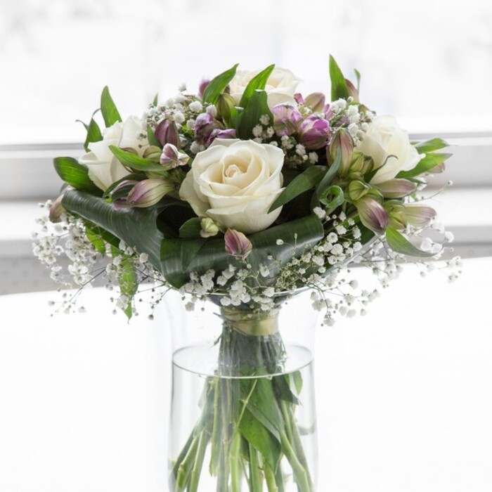 A poised and romantic bouquet of white roses, lavender alstroemeria, and snow-like baby's breath is arranged in a tall, clear glass vase, illuminated by soft natural light that diffuses across a pale background, much like the gentle daylight filtering through a Highgate bay window. The roses are the stars of the composition, generously bloomed and velvety, with ivory petals spiralling outward in flawless concentric layers, creating a sense of purity and timeless grace. Tucked among them are multiple stems of alstroemeria in muted shades of lavender and dusky lilac, some buds tightly closed with green sepals still clasping their tips, others partially open to reveal slender, tapering petals streaked with deeper purple veins. These bring elegant movement and a subtle wash of colour that plays beautifully against the calm white of the roses. Wisps of baby's breath thread between the larger flowers, each tiny white floret contributing to an overall impression of lightness, as though a fine floral mist has settled gently over the arrangement. Surrounding greenery in rich, vibrant tones adds both contrast and structure: broad, dark leaves frame the outer edges, while finer, more delicate foliage curls and weaves within the design. Through the transparent glass of the cylindrical vase, the neatly cut stems are visible, crossing and aligning in clean lines, submerged in crystal-clear water that hints at just-freshly-prepared quality. The composition is rounded and full, yet never heavy, radiating a quiet elegance suited to a condolence message delivered to a home off Highgate Hill, a celebration dinner near Dartmouth Park, or a simple gesture of admiration. It feels like a bouquet crafted by a florist who understands how to blend gentle colour, texture, and emotion into something both soothing and memorable.