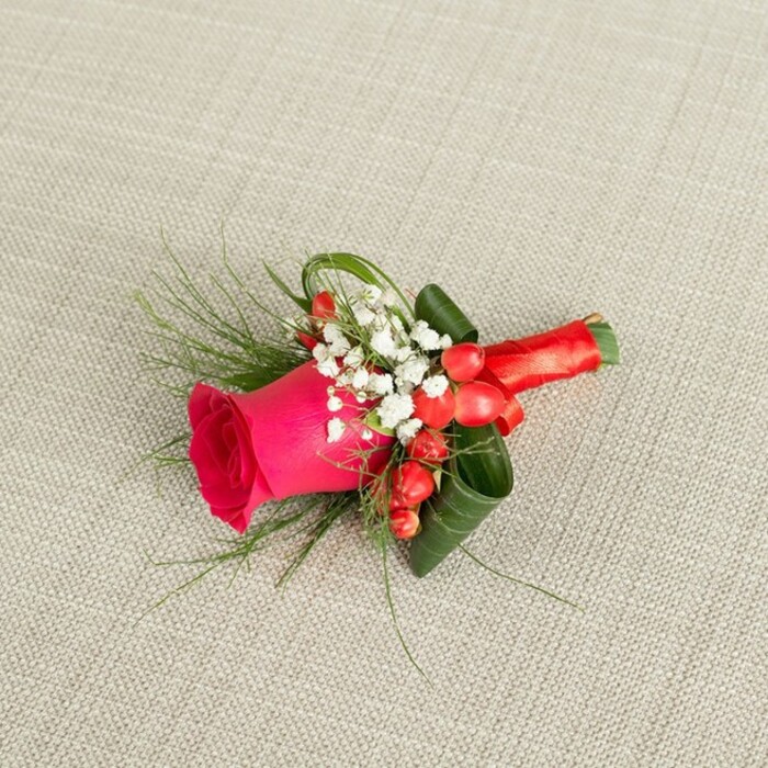 A refined floral boutonniere is laid out diagonally across a light beige, woven fabric backdrop, the gentle texture of the linen-like material giving the impression of a well-loved suit or heirloom tablecloth in a Highgate home. The arrangement runs from the lower left to the upper right, guiding the eye along its elegant line. At the centre of this design sits a striking fuchsia rose, its dense layers of petals tightly cupped yet starting to open, showcasing the flower at an ideal moment of freshness and vibrancy. Around the rose, small clouds of white baby's breath gather, each delicate bloom on fine stems adding a lace-like softness and creating a beautiful contrast against the saturated pink. Interspersed between these white clusters are glossy red hypericum berries, their rounded forms and rich colour echoing the joy and warmth of a celebration, perhaps a wedding taking place near Highgate Wood or a formal event held in one of the area's historic venues. Wispy, fern-like greenery stretches gently outward, lending motion and a sense of natural ease, while darker green leaves are thoughtfully curled and tucked close, shaping the boutonniere into a compact, well-balanced silhouette suitable for a jacket lapel. The stems are firmly wrapped in a glowing red satin ribbon, tied into a tidy knot that feels both secure and decorative, catching the light with a subtle sheen. Photographed from a slightly raised angle, the image highlights each carefully placed element and the harmonious colour palette of pink, white, green, and red. It conveys a mood of romance, care, and quiet sophistication, as though prepared by a florist who understands the intimate moments and local character of Highgate's celebrations.