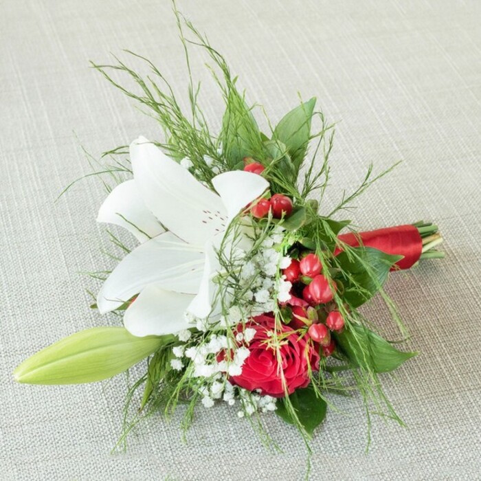 This sophisticated boutonniere or compact bouquet rests serenely upon a pale, textural fabric surface, reminiscent of the charming linens at tea rooms in Highgate Village. Central to the arrangement, a generously open white lily boasts smooth petals adorned with gentle cinnamon speckles and vibrant yellow anthers that catch the soft light filtering through nearby trees. Next to it, a lush, deeply red rose blooms with tightly layered, velvety petals, providing a striking, dramatic contrast. Baby's breath is delicately interspersed throughout, its fine white blossoms softening and brightening the design, while clusters of vivid, glossy red berries add sparkling highlights and bring a festive mood. To one edge, a slender, pale green lily bud peeks out, promising future beauty. The lushness is deepened further by airy, feathered ferns and contrasting dark green leaves, framing the florals in natural elegance. The bouquet's stems are meticulously bound with a shiny red satin ribbon, its texture and tone harmonizing perfectly with the rose and berry accents. Artfully composed by a seasoned Highgate florist, this floral piece-ideal for weddings, anniversaries, or formal gatherings-embodies both warmth and sophistication, echoing the greenery and grandeur of Hampstead Heath and Waterlow Park.