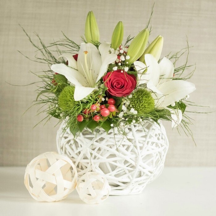 A stylish floral centerpiece has been thoughtfully arranged as if ready for a wedding breakfast or anniversary dinner in a Highgate dining room overlooking the treetops. The flowers are presented in a distinctive, round, white vase crafted from thin, woven strands that cross and loop to form an open, airy lattice. This basket-like construction allows glimpses of stem and shadow, turning the vase into a sculptural feature that enhances the arrangement rather than competing with it. From this elegant base, a generous bouquet of lilies, roses, textured greens, and berries spills naturally upward and outward. Several white lilies dominate the design, some fully open with long, arching petals that curl at the tips, others in smooth, elongated buds still held close. Inside the open blooms, delicate speckles and gently protruding stamens tipped with yellow add nuanced detail and a sense of freshness. Threaded among the lilies are deep red roses, their petals pressed closely together in tight spirals, with one especially eye-catching rose positioned near the centre. Its velvety surface and rich tone infuse the piece with romance and intimacy. Rounded, bright green pom-pom blooms sit at different heights, providing playful texture and a lively, contemporary twist beneath the more classical flowers. Fine sprays of baby's breath float between the stems, their countless tiny white flowers creating a soft haze, while shiny red berries add small, jewel-like accents evoking seasonal celebrations in Highgate Village. A mixture of slender, feathery fronds and broader, darker green leaves fills out the shape, echoing the layered greenery of nearby Waterlow Park. To the left of the vase, two pale wooden decorative spheres with a similar open-weave pattern rest on the surface, reinforcing the cohesive design. The softly textured, neutral background keeps all attention on the flowers, making this arrangement perfect for refined table styling or local delivery in Highgate.