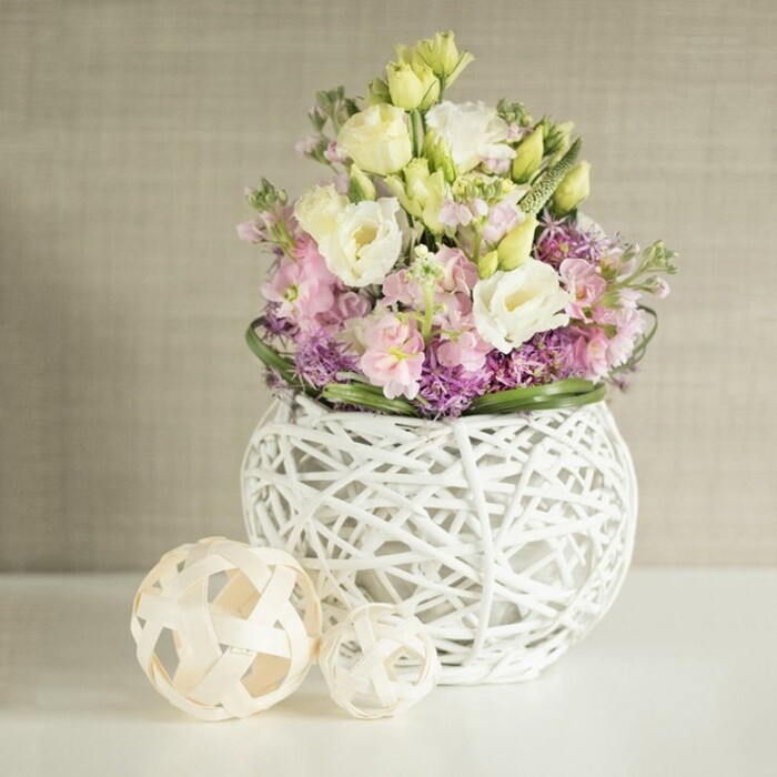 This radiant arrangement encapsulates the refined natural beauty of Highgate with a harmonious display of pastel spring blooms. Creamy white and pale yellow eustoma, beloved for their delicately ruffled, multi-layered petals, take center stage, enveloped by slender sprays of blush pink stocks that impart height and a gentle air of romance synonymous with weddings in Highgate's historic venues. At the base, a carpet of deep purple aster or allium, densely clustered, provides texture and visual intrigue, enhanced by a sculpted border of vivid green leaves that echoes the thriving gardens of Waterlow Park. The bouquet emerges from a distinctive spherical white vase, its surface meticulously woven from painted rattan or fine twigs in an open, basket-like lattice, allowing glimpses of the stems and creating interplay between lightness and abundance. Resting nearby, two pale rattan spheres, each fashioned with an open basket-weave, complement the vase's artisan sensibility and invoke the hand-crafted décor often featured in homes along Highgate Hill. Soft natural light spills across the pristine white surface and highlights the subtle vertical patterning of the beige wall behind, fostering a sense of tranquility and effortless sophistication. This beautifully curated arrangement would grace wedding tables at Kenwood House or add a touch of springtime elegance to any event in Highgate, embodying the care and artistry that Flowers Highgate brings to each and every creation.