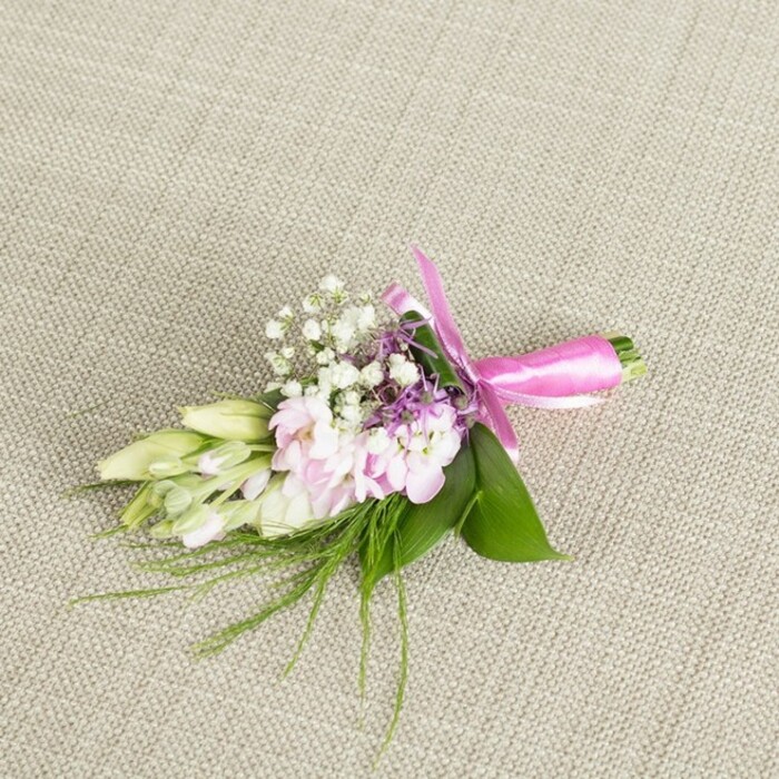 A meticulously arranged boutonniere or small corsage lies centred on a pale greige woven fabric, its linen-like texture visible in soft, criss-crossing threads that evoke the understated elegance of a Highgate boutique. The floral cluster is composed predominantly of soft pink blooms with many delicate, layered petals, resembling miniature stocks or finely ruffled garden flowers that have just been cut at their freshest. Each blossom displays a gentle gradient of colour, from a whisper-light pink at the outer edges to a more concentrated blush nearer the throat of the flower, giving the whole cluster a dreamy, romantic glow. Interlaced with these are airy sprigs of tiny white baby's breath, the minute star-shaped florets creating a cloud-like halo that lightens and softens the composition. Small accents of lavender or dusky purple florets peek through the pastel tones, subtly deepening the colour story and adding interest, much like the shifting tones of evening skies over Highgate Woods. Along one side, a line of elongated buds in creamy white fading to pale green, reminiscent of freesias or similar slender flowers, stretches outward; their closed forms speak of anticipation and blooms about to unfurl. The floral heads rest upon a bed of lush, broad, vibrant green leaves that curve protectively around the stems, their smooth, slightly glossy surfaces catching the light. From beneath, delicate threads of feathery, grass-like greenery splay gently outward, lending a hint of wildness to the polished design. The stems are neatly bound with a striking fuchsia satin ribbon, its bright colour a joyful counterpoint to the soft pastels, and tied into a poised, symmetrical bow with smooth loops and tidy trailing ends. Crafted with the care of an experienced local florist, this piece feels perfectly suited for a Highgate wedding, a prom, or a special celebration, a small but meaningful flourish that photographs beautifully against neutral fabrics.