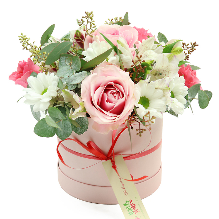 An exquisitely styled floral hatbox arrangement is displayed against a pure white backdrop, its understated luxury echoing the refined feel of Highgate's village boutiques. The container is a cylindrical box in a soft pastel pink, with a velvety, matte finish that appears smooth and tactile. A slim coral-red satin ribbon is wrapped around the lower portion of the box and tied into a small, neat bow at the front, drawing the eye downward to a cream-coloured tag that hangs from the knot. The tag, printed in delicate green script, suggests a personalised note or the mark of a local florist. Above this elegant base, flowers billow over the rim in a lush, dome-shaped bouquet. At its heart is a prominent, fully opened rose with pale pink outer petals deepening into a more saturated pink swirl at the centre, giving a romantic, painterly focal point. Surrounding this hero bloom are clusters of white chrysanthemum or daisy-like flowers, their bright green centres providing a clean, fresh contrast to the soft pinks. Threaded throughout are slender white alstroemeria with trumpet-shaped petals, each bloom lightly speckled with fine markings that add interest without overpowering the composition. Pockets of deeper fuchsia and rich pink blossoms-likely sprigs of spray roses or lisianthus-add intensity and depth, much like vibrant florals spotted in Highgate's hidden gardens. Eucalyptus with its rounded, silver-green leaves emerges between the flowers, accompanied by darker green foliage and fine stems topped with small green buds, building volume and creating a sense of natural movement. The even lighting across the arrangement allows every texture-from velvety petals to satiny ribbon-to be clearly read. This bouquet radiates a soft sense of celebration and affection, making it an ideal choice for birthdays, romantic gestures, or thoughtful thank-yous delivered across Highgate, Archway, and surrounding North London neighbourhoods.