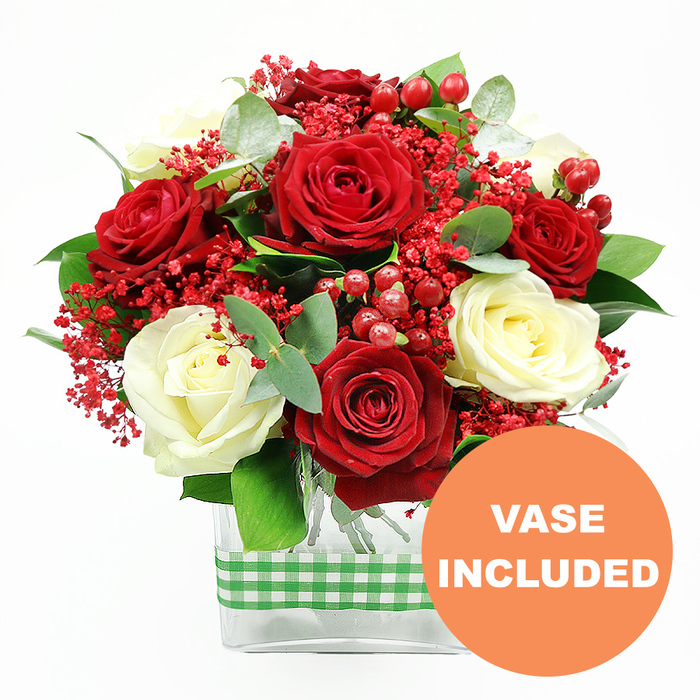 A striking arrangement of roses and seasonal accents is depicted in this image, set against a pure white background that highlights the colours and forms with clarity. Dominating the upper half of the composition, numerous deep red roses and creamy white roses are closely grouped, creating a lush, dome-like mound of petals. Each rose appears carefully selected and placed, with flared outer petals and tighter inner whorls, evoking an impression of fullness and luxury that would complement stylish Highgate interiors and cosy cafés along Highgate High Street. Interwoven through the roses are fine sprays of small, vivid red filler flowers, reminiscent of dyed gypsophila, which lend a delicate, cloud-like quality and soften the contrast between the bold primary blooms. Dotted among the petals, bright red hypericum berries stand out as shiny, rounded accents; their cluster-like formation adds dimension and a sense of festive cheer, subtly suggesting winter celebrations or romantic evenings. Framing the flowers is an abundance of greenery: deep green foliage provides a robust background, while eucalyptus sprigs introduce a cooler, silvery-green hue that hints at fresh, aromatic notes. The stems of all these elements converge neatly into a clear, rectangular glass vase occupying the lower part of the image. The vase's transparent walls reveal the bundled stems lined up in the water, underscoring the design's clean, contemporary style. Around the midpoint of the vase, a green and white gingham ribbon is wrapped snugly, its traditional check pattern bringing an inviting, rustic charm to contrast with the refined roses-much like the blend of village atmosphere and city sophistication found around Highgate Wood. In the lower right corner, a vivid orange circular badge bears the words VASE INCLUDED in bold white letters, making it clear that this bouquet is supplied with its container and arrives ready to display for anniversaries, Valentine's Day, or heartfelt gifts across Highgate.