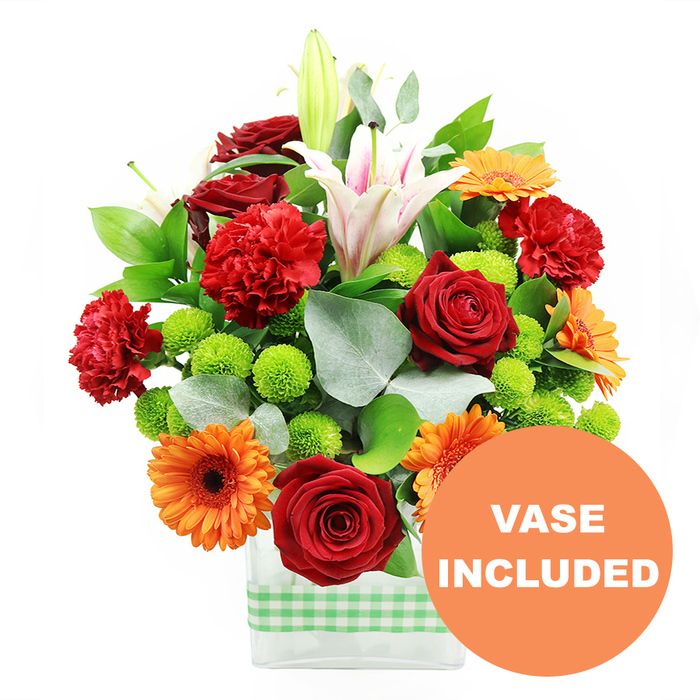 A richly coloured floral display is presented in a low, rectangular clear glass vase, the entire arrangement photographed against a pure white background that makes each flower's hue stand out vividly, like a contemporary centrepiece ready for a Highgate terrace brunch. Around the base of the vase, a light green and white gingham ribbon wraps neatly, its soft checks adding a playful, homely note that contrasts with the vase's clean, modern lines. Above this, the bouquet is dense and lush, filled with an artful balance of colour and texture. Deep crimson roses with velvety, tightly layered petals bring a sense of luxury and romance, while red-orange carnations, with their delicately frilled edges, add volume and a gentle, ruffled softness. Interspersed throughout, bright orange gerbera daisies with dark central discs face outward, their flat petals layered in crisp rings that mimic tiny bursts of sunlight. Nestled among the larger blooms are clusters of lime-green button chrysanthemums, each forming a compact, spherical pom-pom that introduces a lively, almost playful freshness. Rising above the main cluster, slender stems of white oriental lilies create height; some trumpet-shaped flowers are already open, revealing pale pink stripes and golden stamens, while others remain smooth and budded, suggesting fragrance still waiting to unfold. The entire composition is framed by a mix of glossy dark green foliage and sprigs of silvery eucalyptus, whose rounded leaves echo the calm greenery of nearby Highgate cemeteries and parks. In the lower right corner of the image, a bold circular orange graphic partially overlaps the vase and carries the words VASE INCLUDED in clean white capital letters, clearly conveying that the glass container is part of the offering. The mood is bright and celebratory, ideal for birthdays, congratulations, or simply bringing a burst of colour into homes across N6.