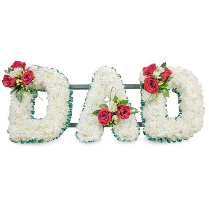 A solemn, handcrafted sympathy piece spelling 'DAD' in large, three-dimensional capital letters, each letter densely covered in a uniform carpet of pure white chrysanthemum blooms. The tiny petals form a soft, cushiony texture that reads as calm and consoling under neutral, diffused light. Each letter is carefully outlined in a ribbed emerald green ribbon, the colour and pleating lending a refined frame to the white faces and sharpening the typographic silhouette. Thoughtfully arranged floral clusters punctuate the design: on the upper left curve of the first 'D' a grouping of rich, deep red roses in various stages of bloom is mixed with small, fragrant white freesia and glossy green foliage; the 'A' bears a corresponding spray near its lower left diagonal, enhanced by a delicate loop of bright green bear grass that lifts the composition; the final 'D' mirrors the first with another composed bouquet on its upper right. Visible between the letters are slender dark metal support rods, subtly tying the three forms into a stable whole without distracting from the flowers. Presented against a crisp white backdrop, every detail-from the velvety rose petals to the feathery chrysanthemum texture and the pleated ribbon edge-reads with clarity. The overall effect is one of dignified tenderness: a personalised floral statement for a father that honours deep affection and steadfast devotion. This arrangement suits a chapel mantel, a crematorium display, or a graveside setting in Highgate or along North Hill where understated craftsmanship and local floral knowledge bring comfort.