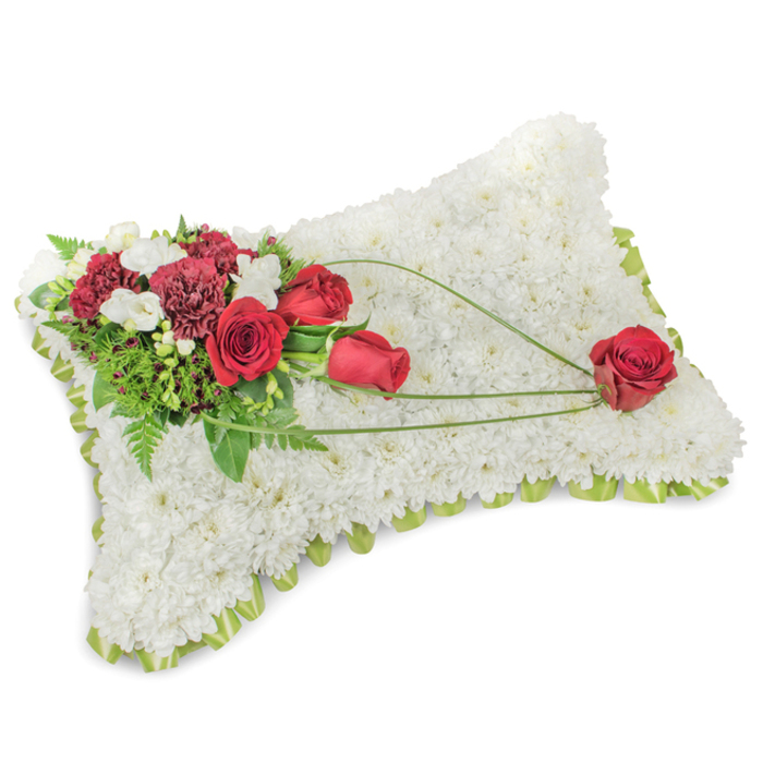 An elegant, rectangular memorial pillow composed entirely of tightly packed white chrysanthemums, their small blooms forming a seamless, tactile surface that suggests peace and purity. The pillow is slightly raised at the centre and edged with a subtle band of light green ribbon or slender foliage that peeks out between the flower heads, giving a neat, finished silhouette against the neutral backdrop. The craftsmanship is precise yet tender, the densely clustered whites creating an effect both restful and refined.

On the upper left a lush floral spray introduces deep, resonant colour: several red roses, some just unfurling and others fully open, are paired with deep burgundy carnations and fine clusters of tiny white accent blooms such as freesias. The mixed textures - the roses' velvety planes, the carnations' ruffled layers, and the little sprays of white - produce a rich, layered focal point. Supporting greenery, including delicate fern fronds and glossy leaves, frames the cluster and adds a natural, garden-fresh backdrop.

From this primary grouping, three slender arcs of bright green bear-grass or similar decorative foliage sweep across the chrysanthemum field toward the bottom right, where one singular red rose rests as a quiet concluding note. The arrangement reads as a tender statement of remembrance, the whites conveying solace while the reds express love and honour. Thoughtfully suited to funeral services, wakes, or commemorative gatherings near Highgate Cemetery, Waterlow Park or the village, this pillow is a classic tribute that balances elegance, scent and sentiment for loved ones and mourners alike.