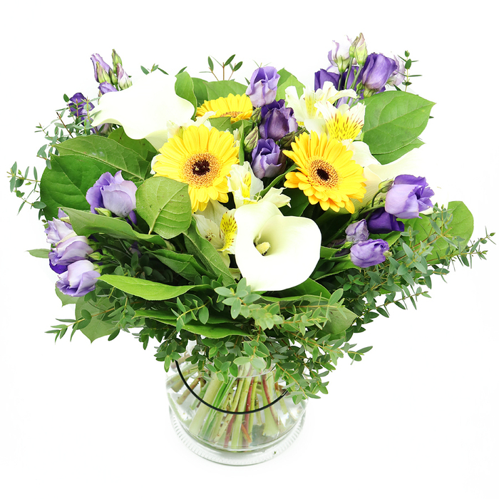 Meticulously arranged in a clear glass cylinder, this mixed bouquet marries joyful yellows with pristine whites and lilac hints in a refined summer palette. The star blooms are several large Gerbera daisies with bright yellow heads and deep centers, radiating warmth and optimism. White Calla lilies rise among them with elegant trumpet forms, lending architectural height. Soft lavender Lisianthus clusters weave through the bouquet, some fully opened, others in bud, providing texture and depth. Smaller white blooms, possibly Alstroemeria with gentle yellow streaks, fill the spaces, lightening the composition. A generous veil of greenery-eucalyptus with its silvery sheen and broad, lush leaves-frames the blooms and keeps the arrangement feeling full and natural. The vase reveals neatly trimmed stems in crystal-clear water, the black tie inside the vase helping secure the design. Against a bright white backdrop, the colours pop with crisp clarity, making this a sophisticated gift for anniversaries, birthdays, or to show appreciation. In Highgate, this design would grace a living room or dining table, and Flowers Highgate offers reliable delivery across Highgate, Muswell Hill, and surrounding streets.