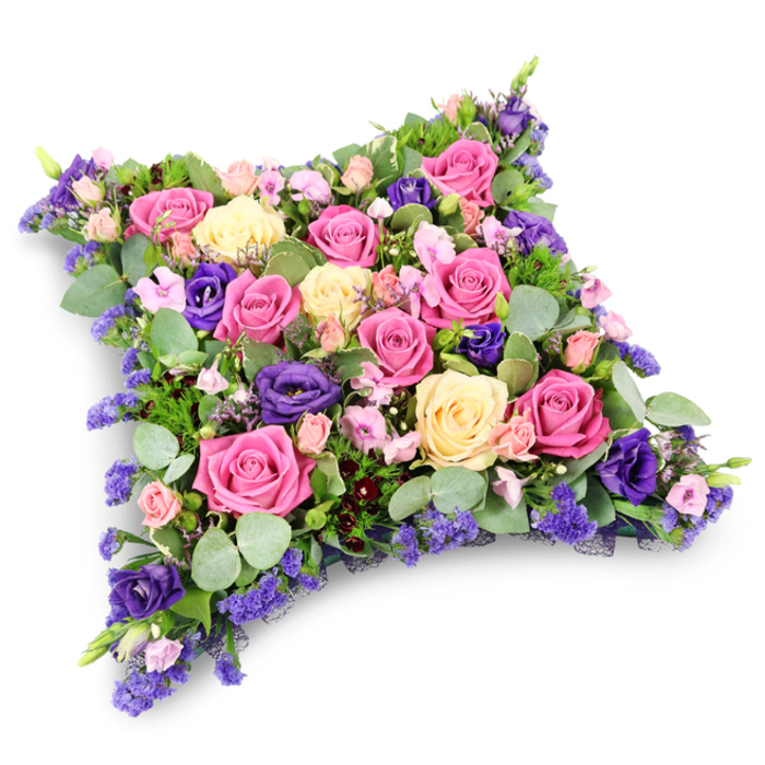 The photograph features an elegant funeral pillow tribute, designed as a cushion-shaped arrangement with a rounded rectangular outline and a gently domed top. The cushion is fully carpeted in flowers, with no visible gaps, creating a lush, almost velour-like surface made entirely of petals and leaves. The main flowers are large-headed roses in vivid fuchsia, delicate pastel pink, and pale buttery cream, each at varying stages of bloom. Some roses are wide open, showing their spiralling hearts, while others are just opening, adding a gentle sense of life and unfolding time. Deep royal-purple lisianthus blooms are nestled among the roses, their soft, ruffled trumpets bringing richness and a slightly cooler, contemplative tone-an echo of the quiet, shaded paths around Highgate Wood and Waterlow Park. Filling the smaller spaces are blush pink spray roses and clusters of tiny lavender and lilac filler flowers, giving the arrangement a fine, intricate layer that invites close attention. Around and beneath the blossoms, an assortment of green foliage provides a steady, natural base: rounded, silvery eucalyptus leaves introduce a misty green-blue hue, while slimmer, fern-like greenery adds lightness, depth, and shape. Against the plain white background, the cushion appears serene and dignified, hovering visually as if placed on a table of honour before a service. The combination of gentle pastels with deeper purples creates an atmosphere of comfort, remembrance, and quiet love, making it particularly fitting for funerals and memorials within the Highgate area. It is the kind of floral tribute families might send to accompany a casket or urn, symbolising a soft, final resting pillow offered in affection and respect, each stem placed with the care and understanding of a local, sympathetic florist.