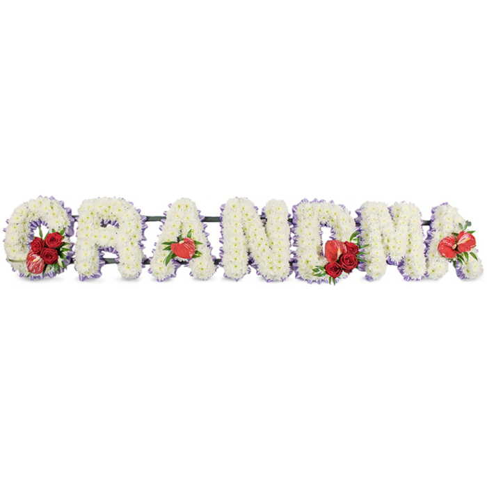 A lovingly detailed floral tribute forms the word GRANDMA in bold, three-dimensional capital letters, the kind of arrangement you might find resting in a tranquil Highgate church or natural burial ground. Each letter is densely covered in immaculate white cushion chrysanthemums, whose compact, button-like blooms overlap to create a soft, cushiony texture with no visible gaps, suggesting care and completeness. Around the perimeter of each character runs a fine band of lilac or light purple ribbon and backing, providing a delicate contrast and outlining the shapes so that the letters stand out clearly against the smooth white background. The florist has placed vivid red accents with deliberate intention along the dark green structural frame beneath the flowers. At the beginning of the word, the G holds a sumptuous cluster of deep red roses, nestled with a single red-and-white variegated calla lily that curves elegantly alongside the white blooms. The first A is graced by one heart-shaped red anthurium, its glossy surface catching the light and echoing the idea of a loving heart. In the centre, the M is similarly adorned with a grouping of velvety red roses and another red anthurium, forming a rich focal point that anchors the design. At the far end, the last A displays one more red anthurium, mirroring the earlier detail and completing the gentle rhythm of colour. The arrangement feels both traditional and deeply personal, ideal for a funeral, wake, or graveside tribute in Highgate or nearby areas such as Dartmouth Park, offering a clear, floral expression of gratitude and remembrance for a cherished grandmother.