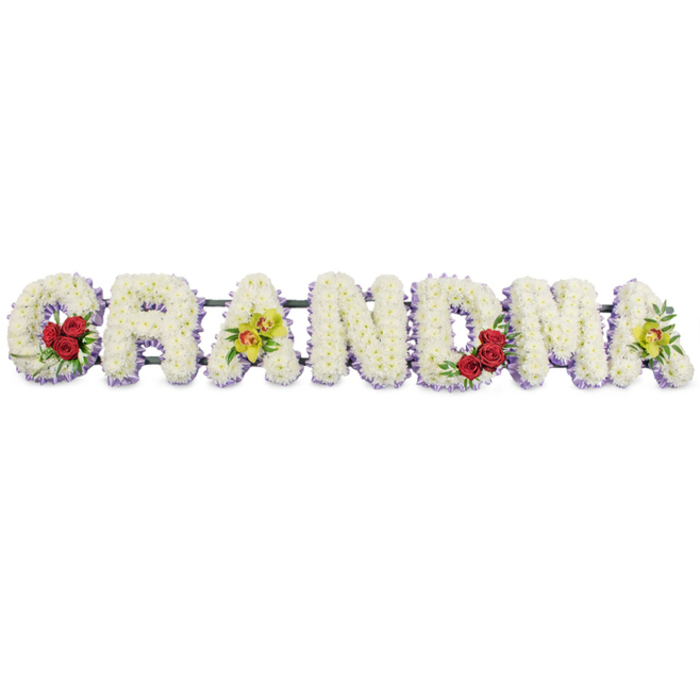 A lovingly composed funeral flower arrangement spells out GRANDMA in bold, uppercase letters, arranged horizontally and photographed against a simple, uncluttered white background that brings to mind the calm light inside churches around Highgate Village. Each letter is meticulously formed from a mass of small, delicate white chrysanthemums or similar blooms, pressed tightly together so that no gaps show, creating a smooth, tactile surface where each tiny petal overlaps the next. Just beneath this blanket of white, a narrow edging of soft lavender or pale purple flowers runs along the base of every letter, adding depth and a faint wash of colour that helps define the outlines, rather like the soft shadows cast by trees around nearby Highgate Wood. The G carries a cluster of three deep red roses at its lower left curve, their velvety crimson petals framed by dark green foliage, a clear focal point symbolising devotion and memory. The first A from the left is distinguished by two yellow-green orchids with intricate reddish-brown centres, placed along the crossbar in a way that introduces an unexpected yet harmonious touch of exotic grace. The R, N, and D remain pure and unembellished in white with their lavender border, giving the central section of the tribute a quiet, contemplative feel. On the M, a second grouping of three deep red roses sits within the lower right, balancing the G and drawing the eye towards the heart of the word. The final A mirrors the first with two more yellow-green orchids, ensuring the composition feels complete and thoughtfully symmetrical. All the letters are fixed onto a discreet dark green or black metal frame that links them along the back, providing structural support while remaining virtually unseen. This GRANDMA floral tribute is a traditional and deeply personal piece of sympathy floristry, perfectly suited to funeral services, memorial gatherings, or graveside tributes in Highgate.