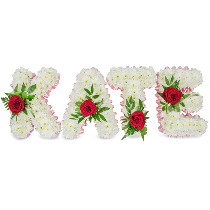 This image presents a large, personalised floral tribute forming the name KATE in block letters, handcrafted from an abundant mass of crisp white chrysanthemums whose tightly packed petals give each letter a rounded, clouded volume. A delicate pale pink ribbon traces the outer contour of every letter, its subtle sheen and soft pleating lending the arrangement an understated, refined edge that frames the white blooms. Each letter carries a single accent of intense, ruby-red rose-deep, velvety and visually commanding-tucked into a generous cluster of fresh green foliage comprising broad, glossy leaves and finer fern fronds that provide both support and a lively textural counterpoint. The placement of these red focal roses is carefully considered: the rose on the K is located on the lower portion of the left vertical stroke, the A's bloom crowns the apex of its triangular form, the T's rose nestles at the left base of the horizontal crossbar, and the E's flower sits on the middle bar, creating a harmonious visual rhythm across the name. Photographed on a pristine white background, the arrangement conveys a classic, respectful expression-white for purity, red for love, green for continuity-suitable for funeral chapels, graveside tributes, or memorial displays. As a florist familiar with Highgate Village customs and services around the Pond and North Hill, I recognise how this piece reads in a room: calm, thoughtfully composed, and quietly consoling to those saying goodbye.
