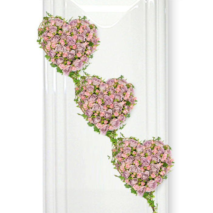 The image showcases a refined triple-heart funeral arrangement that seems perfectly at home in Highgate's blend of old-world charm and contemporary elegance. Three distinct hearts, each sculpted entirely from roses, flow diagonally from the top left corner to the bottom right, creating a soft, cascading effect similar to looking down a gentle slope towards Highgate Cemetery's winding paths. Every heart is densely filled with rose blooms in muted shades of lavender and mauve, interspersed with smaller, pastel pink roses that lighten and animate the design. The petals overlap to form a thick, velvety layer, their edges curling and folding in natural, intricate patterns that speak of careful craftsmanship. No large gaps of greenery show through the blooms; instead, the roses create the impression of a continuous floral tapestry. Surrounding and linking the hearts are flexible threads of fresh ivy, their glossy green leaves catching the light as they frame each heart and run between them. This ivy not only defines the heart shapes but also suggests an unbroken thread of love and remembrance, much like the connections between families who have lived around Highgate Hill and Pond Square for generations. The background is an uncluttered, brilliant white, lit gently so that the shadows are soft and the pastel tones of the roses stand out clearly. On the left side, faint vertical, translucent streaks hint at glass or a reflective interior surface, adding a layer of quiet sophistication. The overall impression is one of romantic dignity and heartfelt sentiment, making this arrangement suitable for a funeral, memorial, or celebration of life where the family wishes to convey deep affection, unity, and ongoing connection beyond the day of farewell.