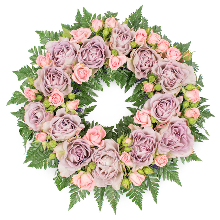 A lush, circular sympathy wreath is seen from above, set against a smooth white background that lets every detail of the floristry stand out, much like a carefully arranged tribute resting in a Highgate chapel. The wreath's structure is flawlessly round, suggesting thoughtful planning and skilled hands. Large, open roses in gentle tones of dusty lavender, antique mauve, and soft greyish-purple provide the primary visual focus, each bloom appearing at its prime, neither too tight nor fading. Their many layers of petals, slightly curled and overlapping, create intricate patterns and velvety textures that draw the viewer's attention from one flower to the next. Interspersed among these larger roses are clusters of diminutive blush pink spray roses and buds, their smaller size and paler tones adding lightness and romantic charm. Some buds show just a blush of pink at the tip, as though on the verge of opening, bringing a sense of quiet anticipation and continuity. The flowers are cradled in a dense collar of dark green fern fronds, whose narrow, finely serrated leaves overlap into a thick, textured border. The fern's deep green contrasts beautifully with the softer floral palette, giving the wreath a grounded, natural presence, as if inspired by the leafy walks around Waterlow Park or the wooded paths near Highgate Wood. Subtle additions of darker foliage or tiny seed heads tucked within the design add depth and shadow, enhancing the wreath's three-dimensional form. The overall composition is calm, balanced, and deeply respectful, perfectly suited to a funeral, cremation, or celebration of life, and reflective of the considered elegance often sought by families in the Highgate community.