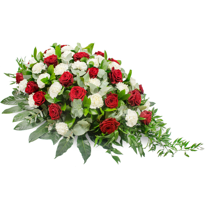 This image shows an elegant, horizontal funeral spray set against a plain white background, allowing the colours and textures of the flowers to speak clearly. The arrangement has an asymmetrical teardrop profile, fuller and more rounded at the left before gradually narrowing into a fine, trailing tip on the right, much like the shape commonly seen gracing caskets in Highgate's historic chapels. Deep crimson red roses are scattered purposefully across the spray, their petals compact and velvety, each bloom acting as a focal point of emotion and depth. Surrounding and softening these roses, numerous white carnations expand in layered, ruffled clusters, creating a cushiony texture that feels gentle and consoling. The interplay of the dark, passionate red with the calm, immaculate white establishes a visual language of love, loss, and serene remembrance. The flowers are embedded in a lush bed of foliage: broad, dark green leaves provide a strong, almost architectural base, framing the arrangement and supporting its elongated form. Among them, softer, silvery-green eucalyptus leaves peek through, lending subtle colour variation and a slightly hazy, romantic quality. Towards the right-hand tip, slender strands of trailing greenery extend outward, conveying a sense of natural movement, as if the spray had just been set down after being carried along Highgate Hill. The overall effect is one of poised, quiet beauty, designed to lie flat on a casket or memorial table. This is the kind of arrangement families in Highgate and the surrounding North London areas might choose when they wish to honour a loved one with a traditional yet deeply heartfelt floral tribute.